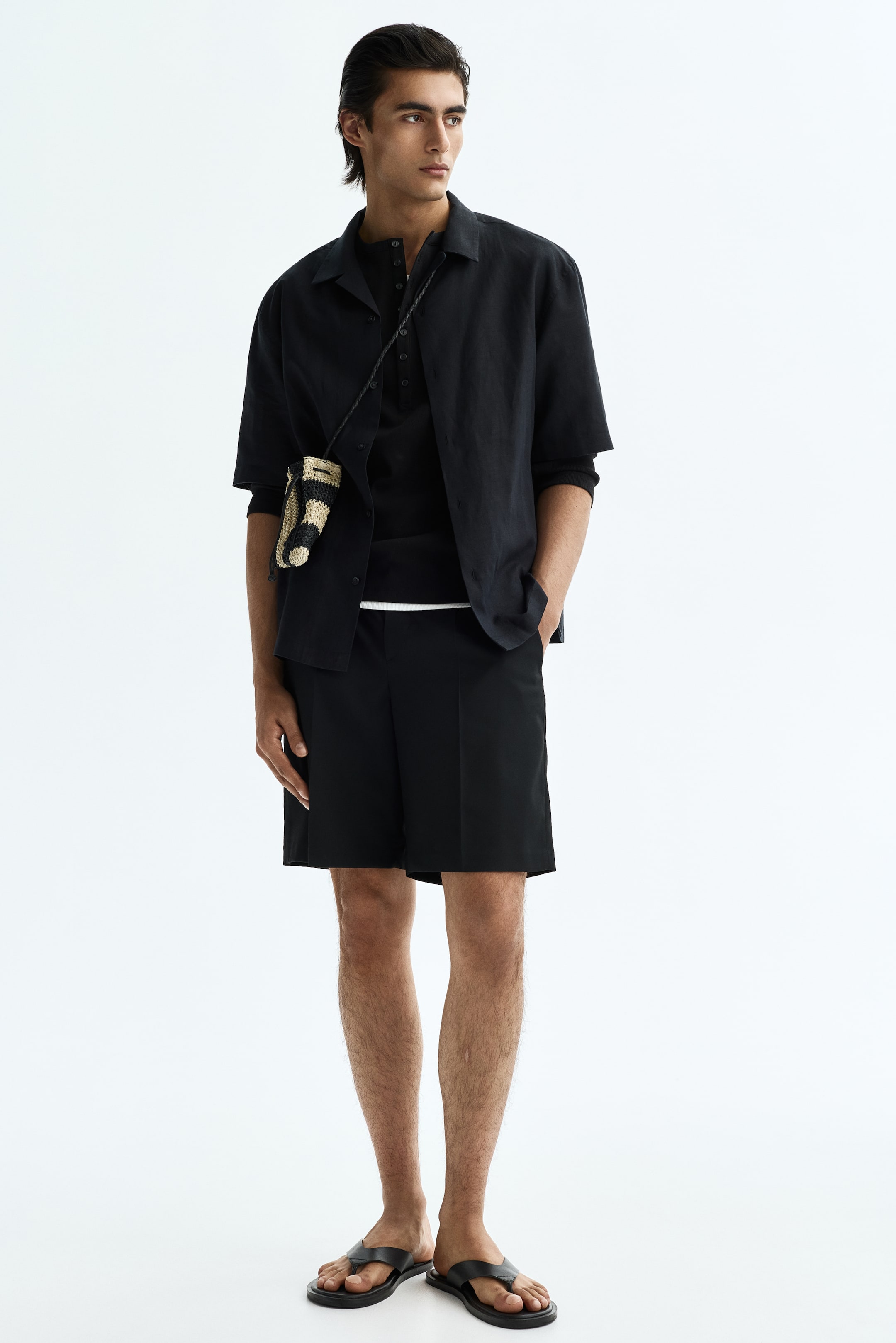 הצגת תמונה גדולה יותר: חולצה שחורה עם שרוולים קצרים וצווארון נלבשת פתוחה מעל חולצת הנלי שחורה עם שרוולים ארוכים, מעוצבת עם מכנסיים קצרים באורך הברך בצבע שחור וסנדלי אצבע שחורים. תיק קטן ארוג עם פסים נישא על המותן של הלובש.