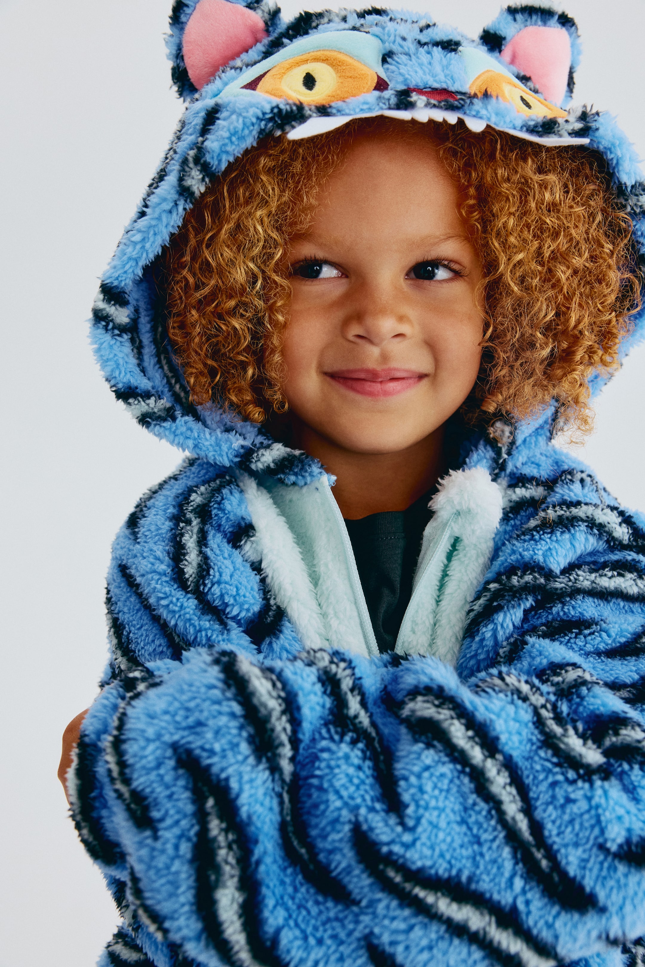 Ver imagem maior: Uma peça de roupa polar com capuz, inspirada num tigre azul, apresenta riscas azuis e pretas numa textura fofa. O capuz foi concebido com orelhas forradas a cor-de-rosa, olhos amarelos e laranja, e dentes brancos, e é visível um forro polar azul-claro לאורך a frente aberta, revelando uma t-shirt verde-escura por baixo.