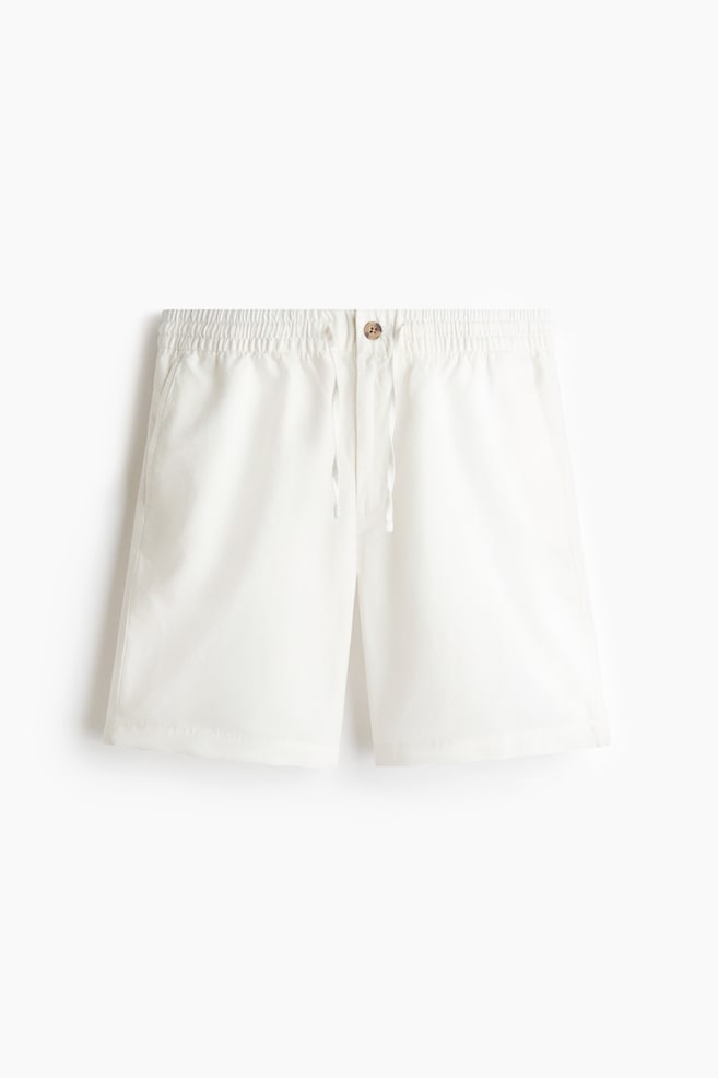 Shorts blancos confeccionados en una mezcla de algodón y lino texturizado se presentan desde el frente, mostrando una cintura elástica con cordón y cierre de botón, junto con bolsillos en las costuras laterales. - 1