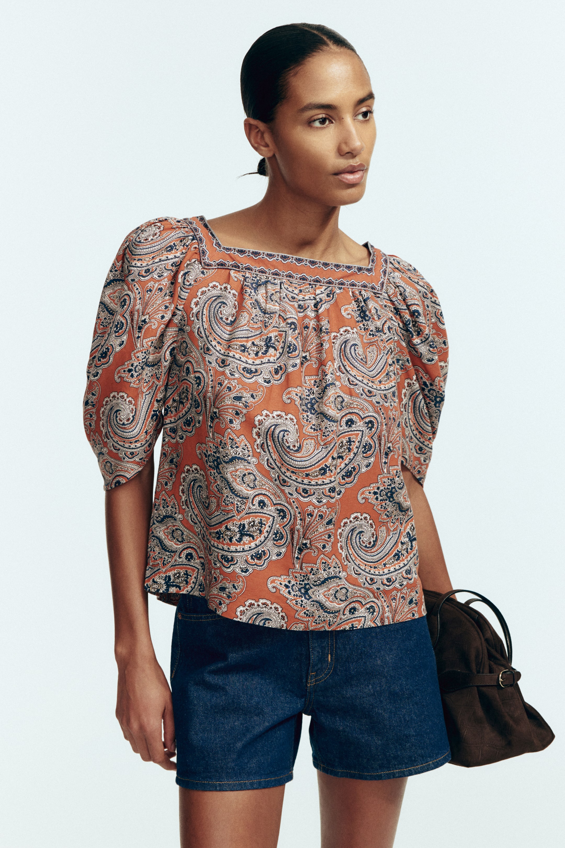 View larger image: A square-neck blouse, angled slightly to the front-right, is presenting an orange-brown base with a detailed blue, white, and light orange paisley print. 