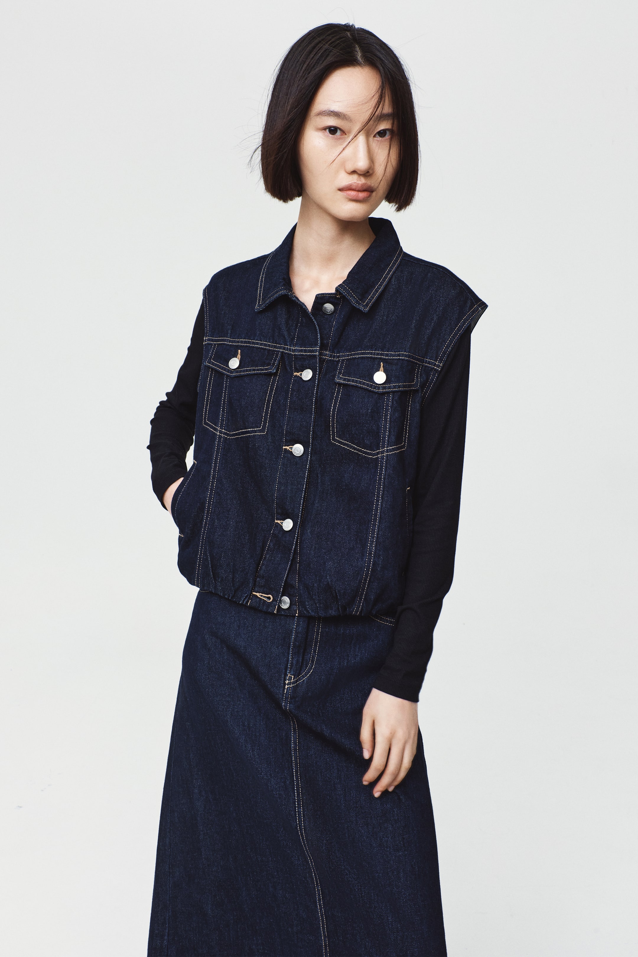 View larger image: A dark blue denim vest featuring a collar, button-up front, patch chest pockets with buttoned flaps, and an elasticated hem is worn by the model. The vest is layered over a black long-sleeve top and paired with a matching denim skirt, with the model placing one hand inside a side welt pocket.