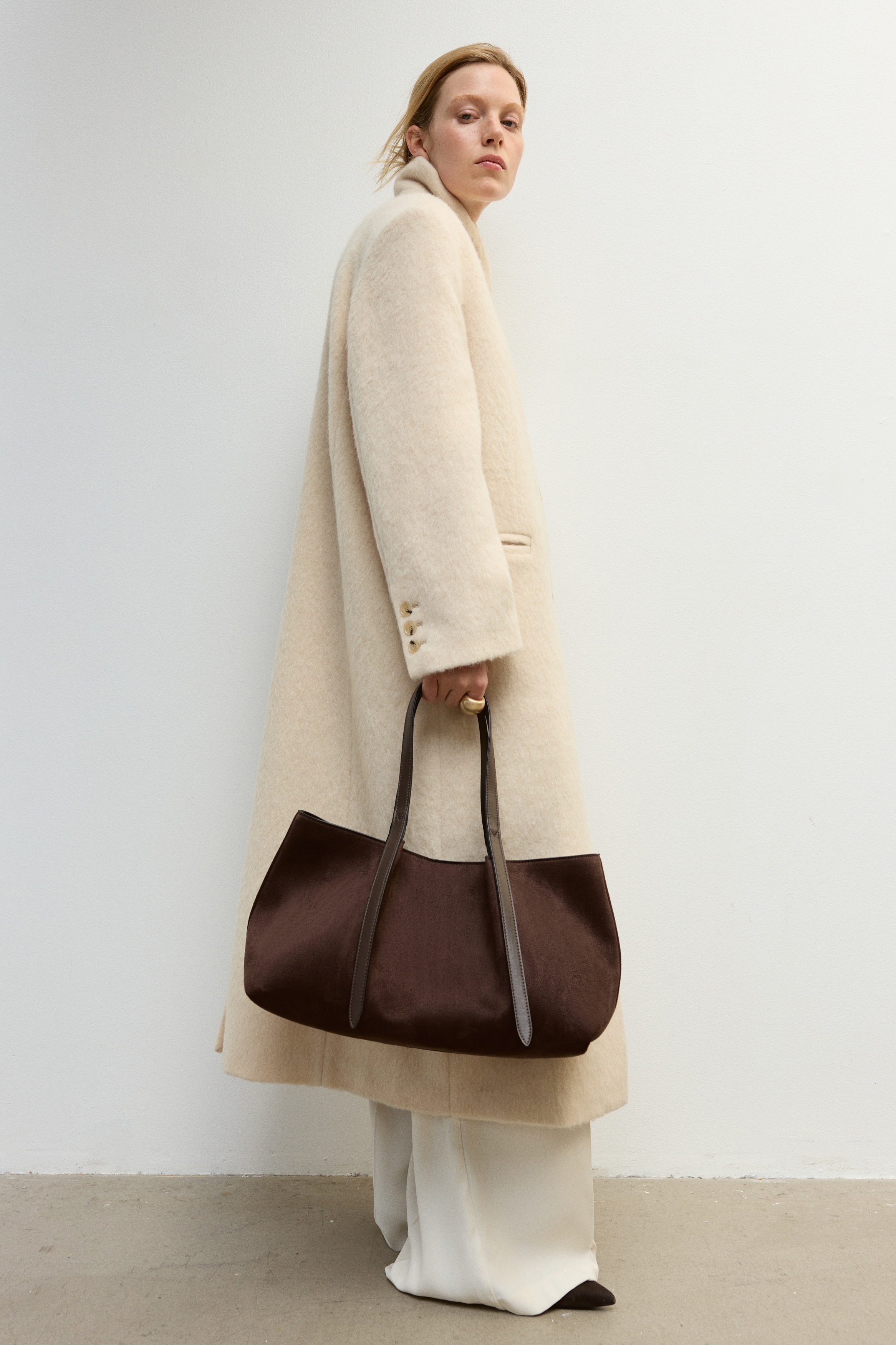 A dark brown suede tote bag is shown in a front view, highlighting a slouchy, unstructured silhouette. The bag features long, flat leather shoulder straps that extend down the front face with loose ends.