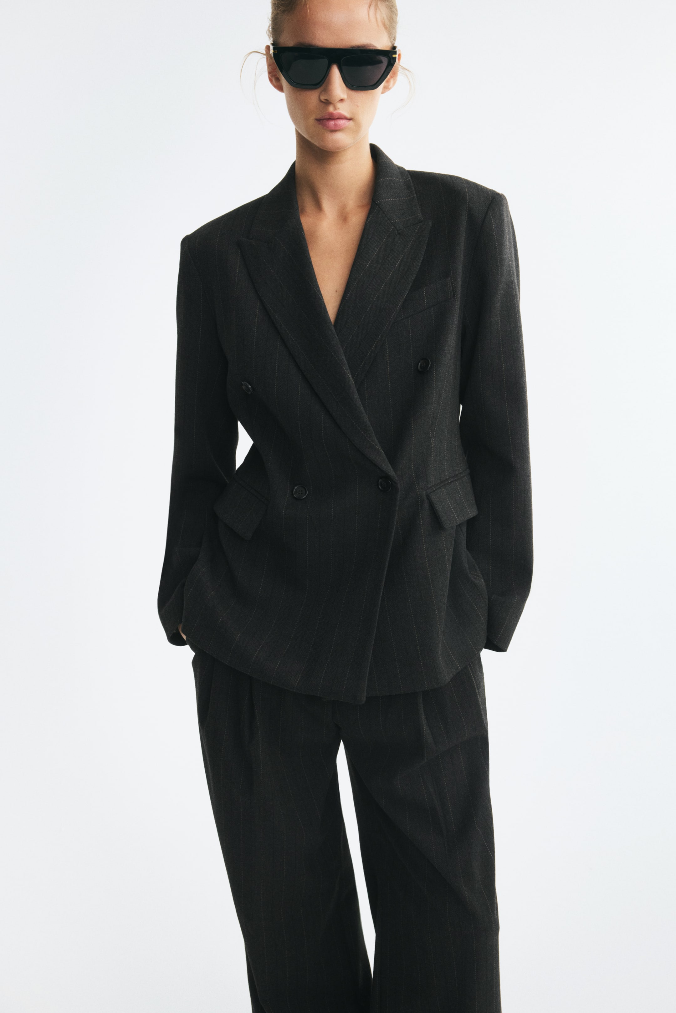 View larger image: A dark grey, pinstriped, double-breasted blazer featuring peak lapels, shoulder pads, and front flap pockets is worn by the model. The fitted blazer is styled with matching pinstripe trousers.