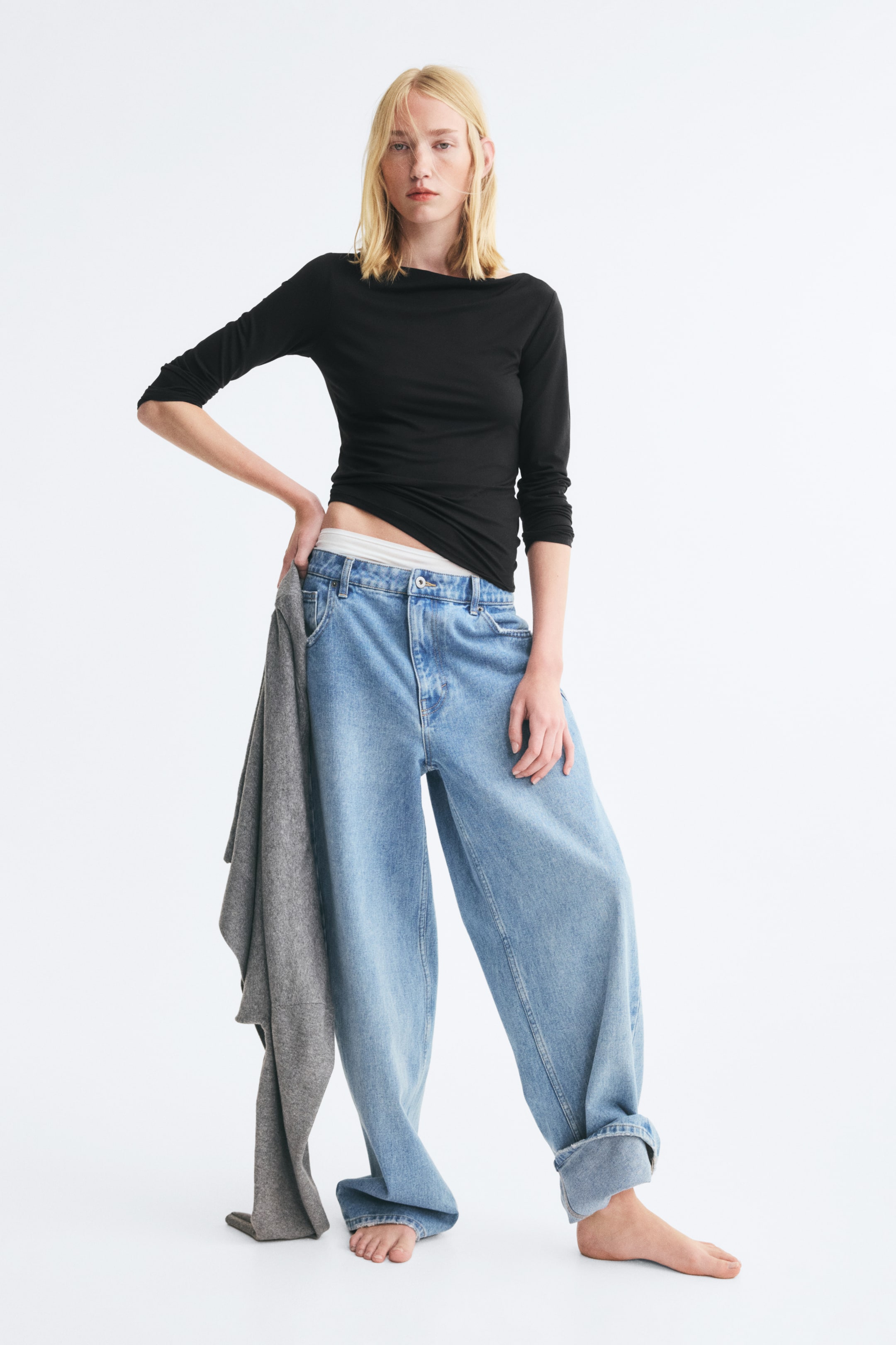 Büyük resmi görüntüle: Model üzerinde, kayık yaka ve uzun kollu, esnek jarse kumaştan oturan siyah bir üst yer alıyor. Üst, geniş paça mavi kot pantolonla kombinlenmiş ve modelin elinde gri bir giysi tutuluyor.