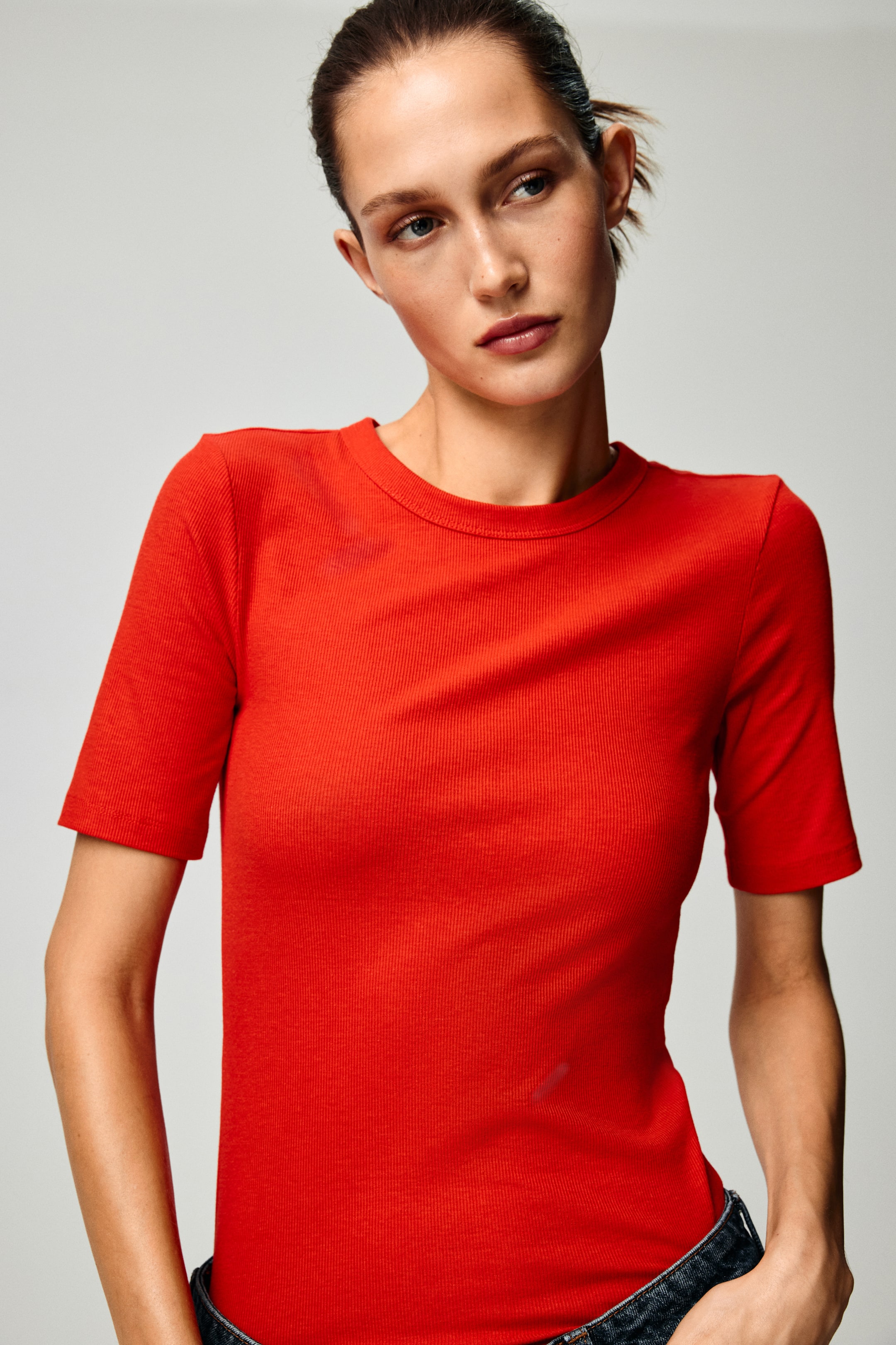 View larger image: A red ribbed-knit T-shirt is worn, featuring a crew neck and short sleeves. It presents a fitted silhouette, with the hem tucked into dark jeans.