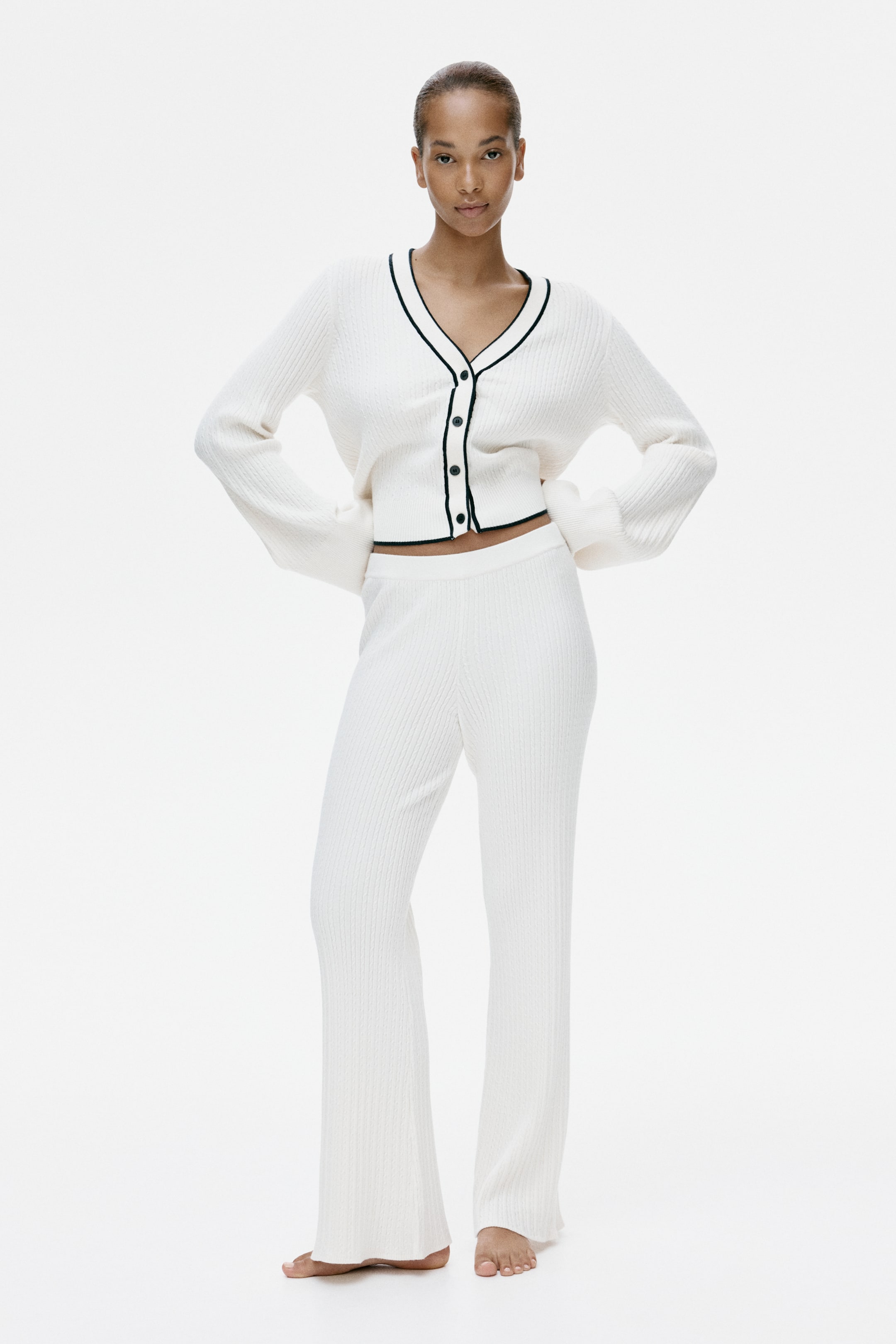 View larger image: A cream ribbed knit set features a cropped cardigan and matching flared trousers, shown front-on. The cardigan has a V-neck with black trim along the neckline, button placket and hem, and voluminous long sleeves. The matching trousers have a relaxed, full-length, wide-leg fit.