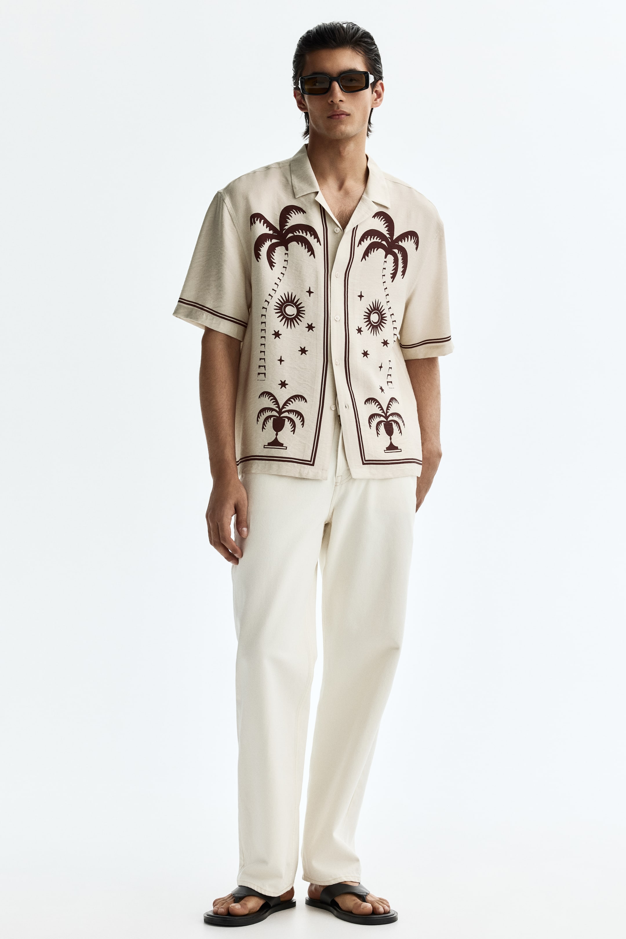 Voir une image plus grande: Une chemise crème à manches courtes présente un col à revers et un motif brun foncé de palmiers, soleils, lunes et étoiles. Elle est dotée d'une fermeture à boutons sur le devant et d'une bande unique accentuant les manches courtes, orientée vers l'avant.