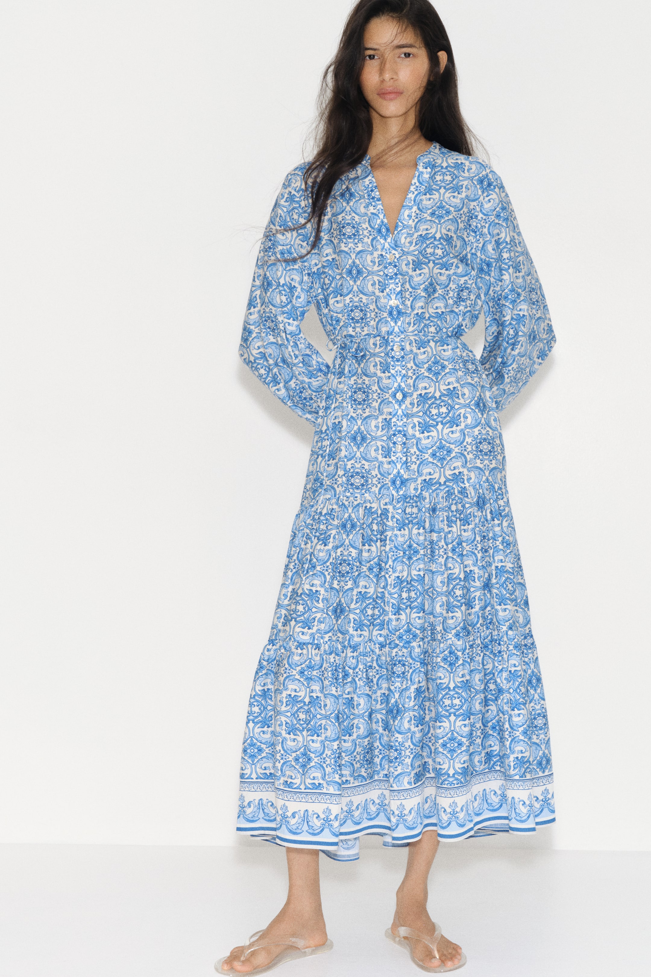 Ampliar la imagen: La modelo lleva un vestido hasta el tobillo con un estampado integral en azul y blanco. Presenta un cuello tipo banda con una abertura en forma de V, botones frontales, mangas largas abullonadas con puños elásticos y un cinturón de lazo tipo espagueti que ajusta la cintura sobre una falda acampanada con volantes recogidos.