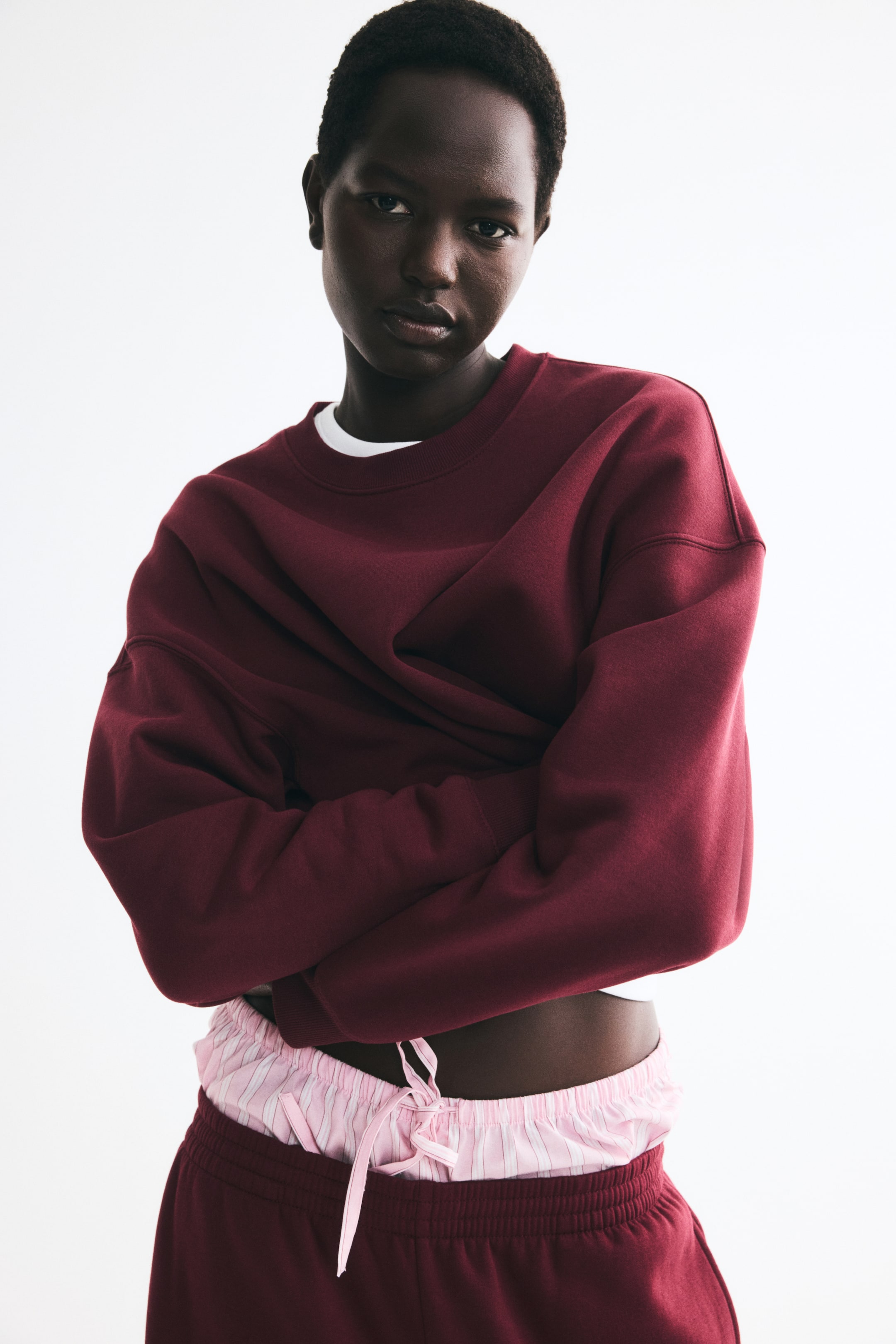 Grotere afbeelding bekijken: Een oversized donkerrode sweatshirt met een geribbelde ronde hals, verlaagde schoudernaden en lange mouwen wordt gedragen door het model, dat de armen over elkaar heeft. De sweatshirt is gelaagd over een wit T-shirt en gestyled met roze gestreepte boxershorts en bijpassende donkerrode sweatpants.