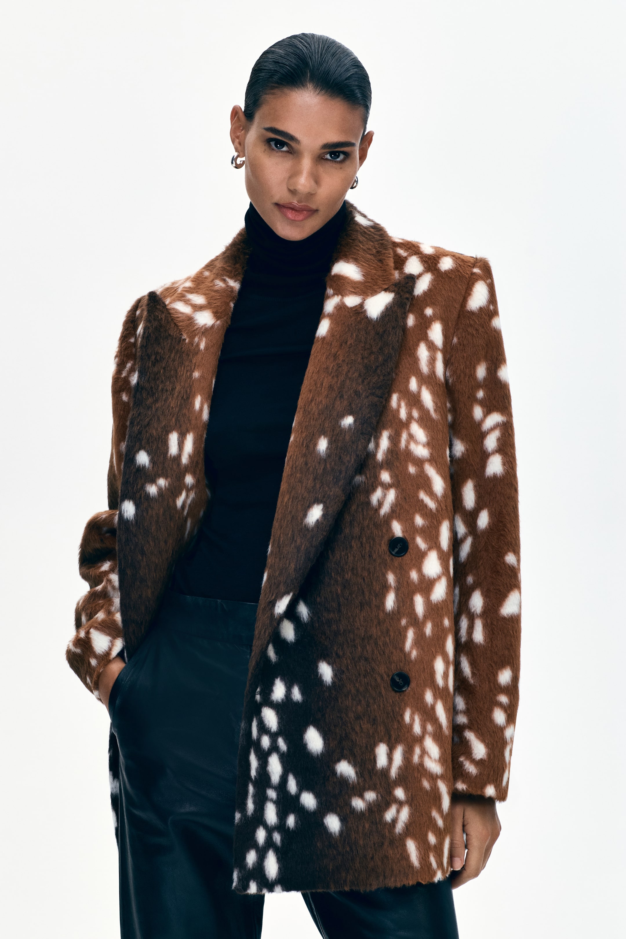 View larger image: A brown and white fawn print faux fur coat, featuring notch lapels and a double-breasted front with two dark buttons, is layered over a black skivvy and black trousers.