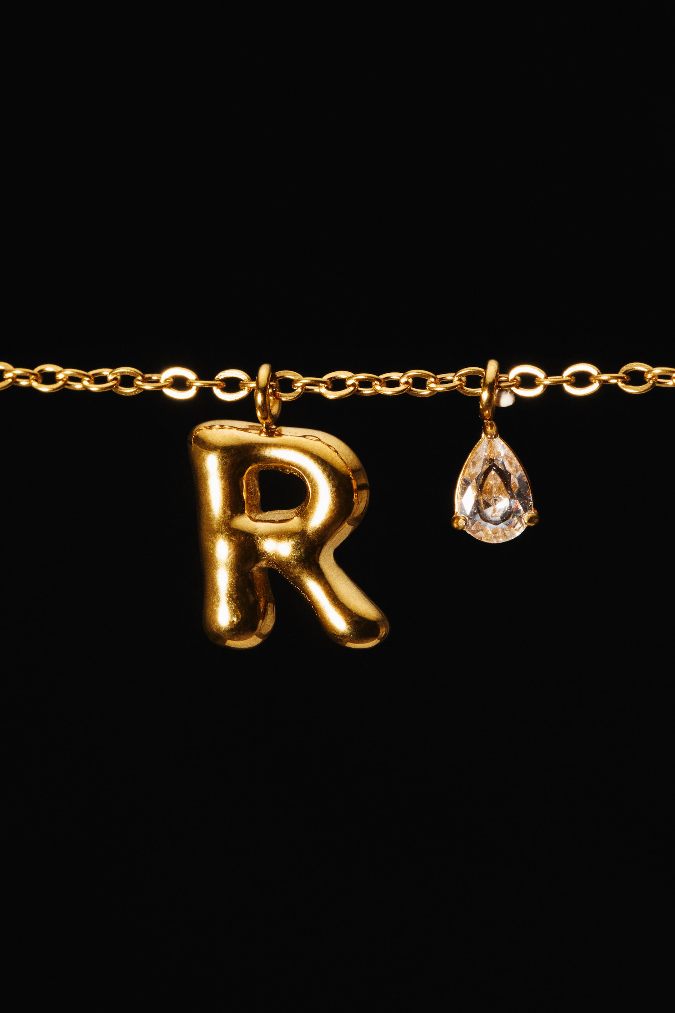 View larger image: A gold-toned stainless steel chain necklace is shown from the front, featuring a puffed letter "R" pendant alongside a small, teardrop-shaped glass rhinestone pendant.