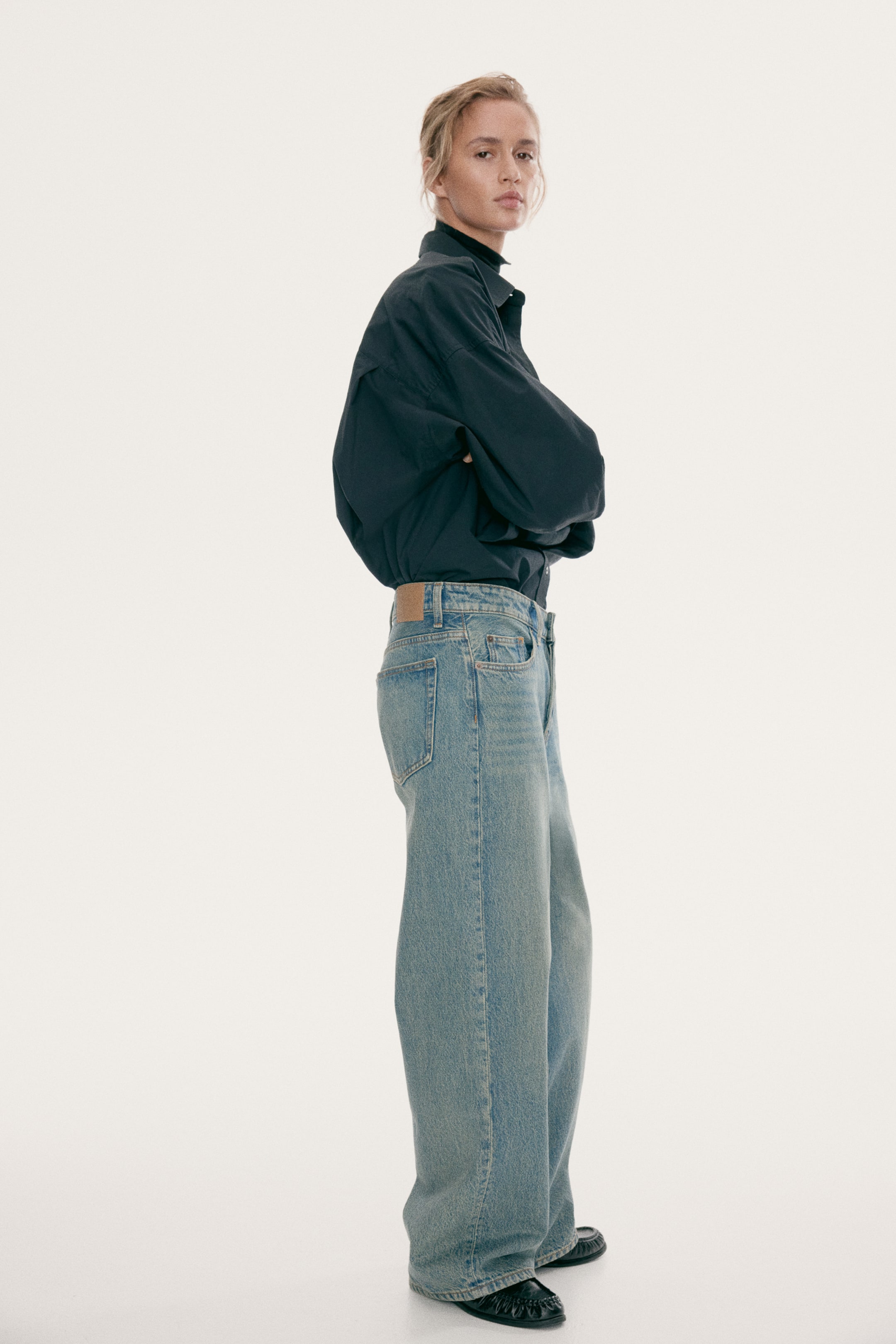 Visa större bild: Ljusblåtvättade jeans med låg midja och lös, vid benvidd bärs av modellen. Jeans med fem fickor kombineras med en instoppad mörkgrå skjorta med knappar och svarta skor, vilket framhäver den baggy siluetten från låret till fållen.