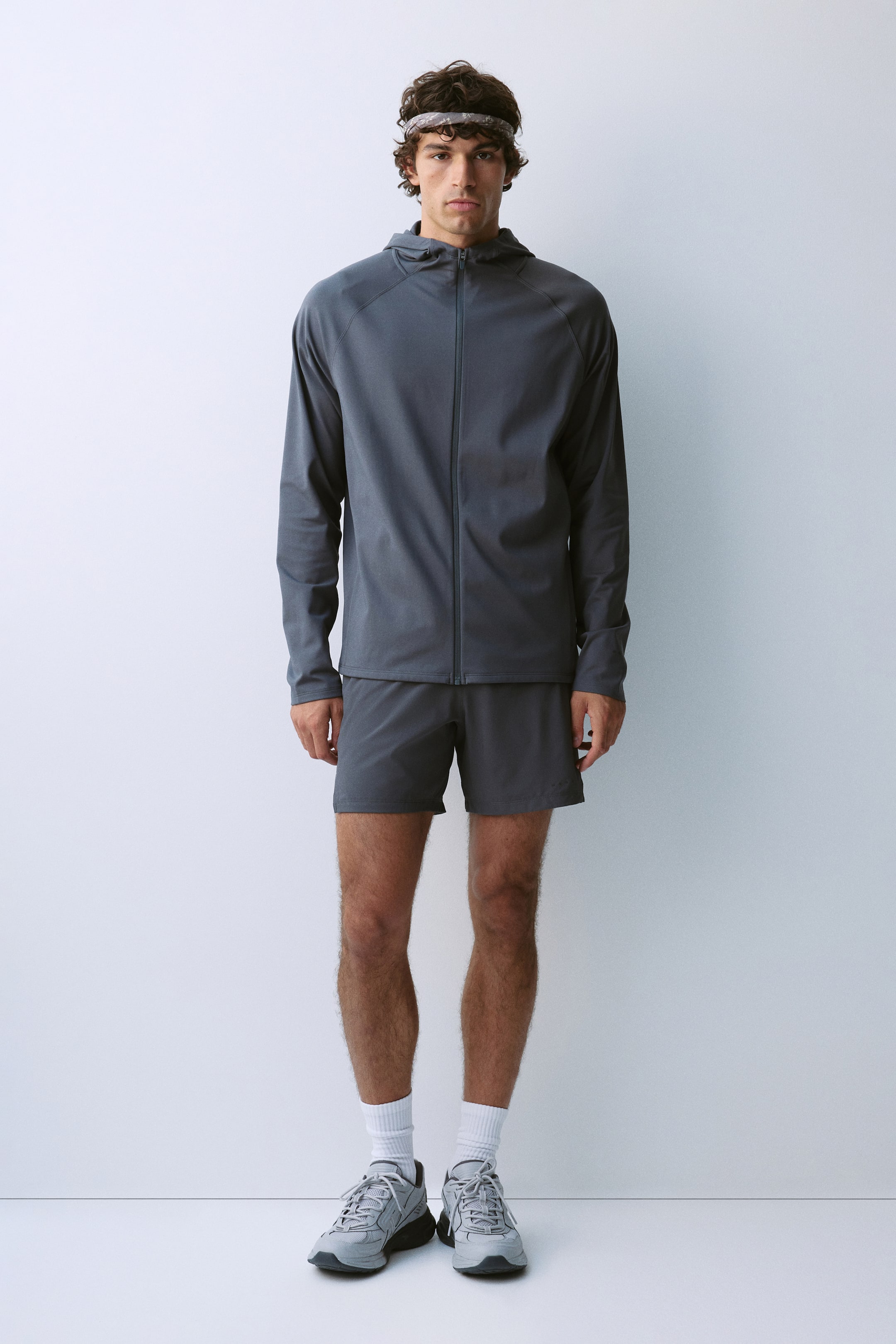 View larger image: A dark grey athletic set consisting of a long-sleeve hooded jacket and matching shorts is being worn by the model. The zip-up jacket features raglan sleeves and is paired with a grey headband, white crew socks, and grey running shoes.