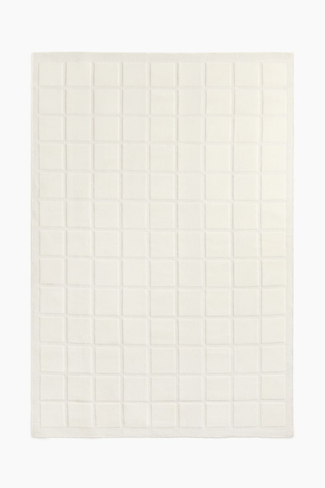 A cream-coloured blanket is lying flat, displaying a grid of raised square patterns across its surface and a subtle ribbed border. - 1