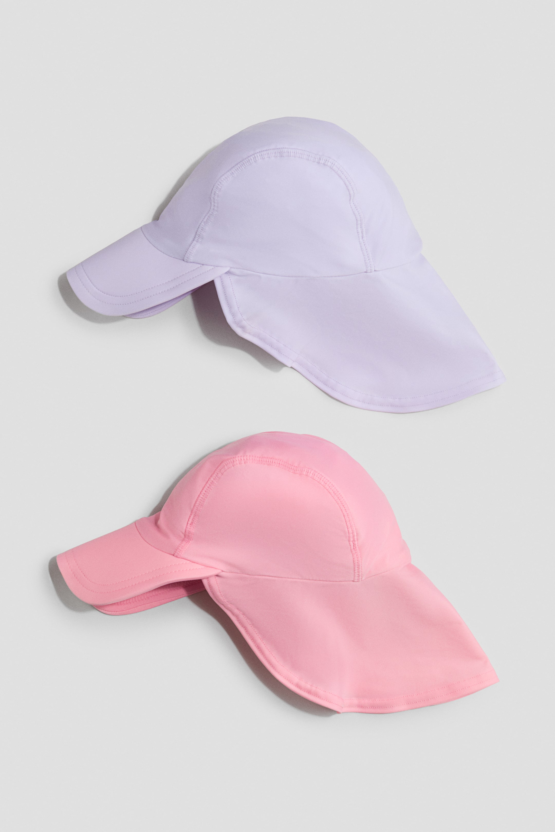 View larger image: Two sun hats, one in light purple and the other in pink, feature front brims, back flaps, and visible flatlock seams.