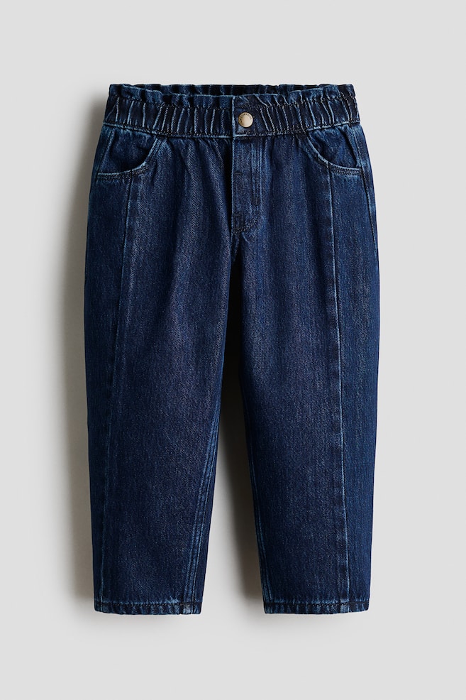 Donkerblauwe denim broek staat naar voren gericht, met een elastische, geplooide tailleband en een enkele messingkleurige knoopsluiting. Ze hebben twee schuine voorzakken en een verticale naad die over het midden van elke broekspijp loopt. - 1