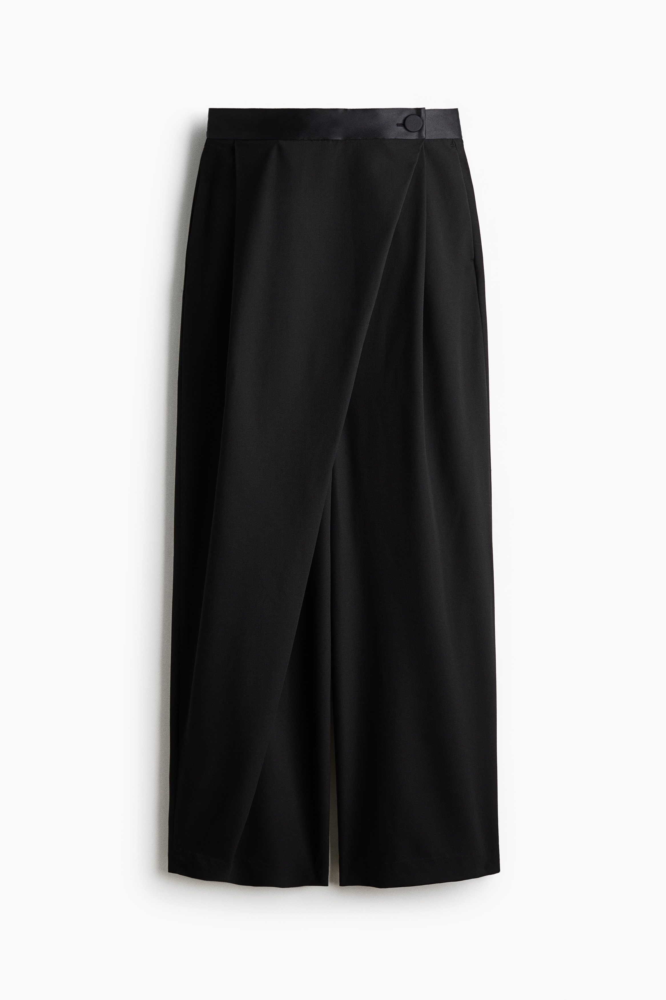 Black wide-leg twill pants featuring a satin waistband, front pleats, and a wrap-front design are being worn by the model. The pants are styled with a coordinating cropped black blazer, a black crop top, and white woven shoes.