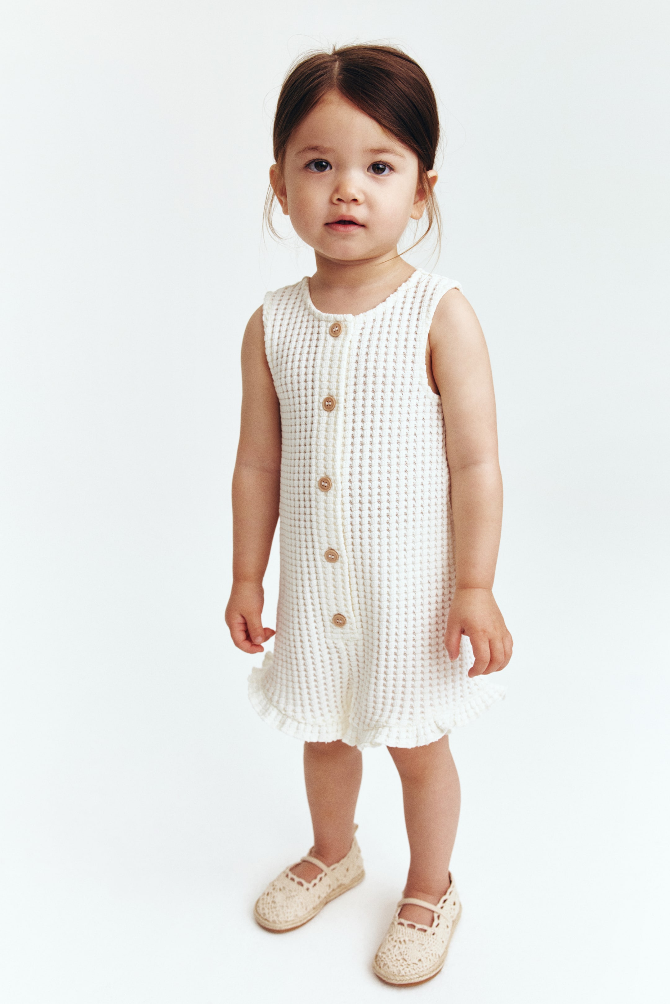 View larger image: An off-white sleeveless waffle knit romper featuring a round neckline, a front button placket with light brown buttons, and ruffled short leg hems.