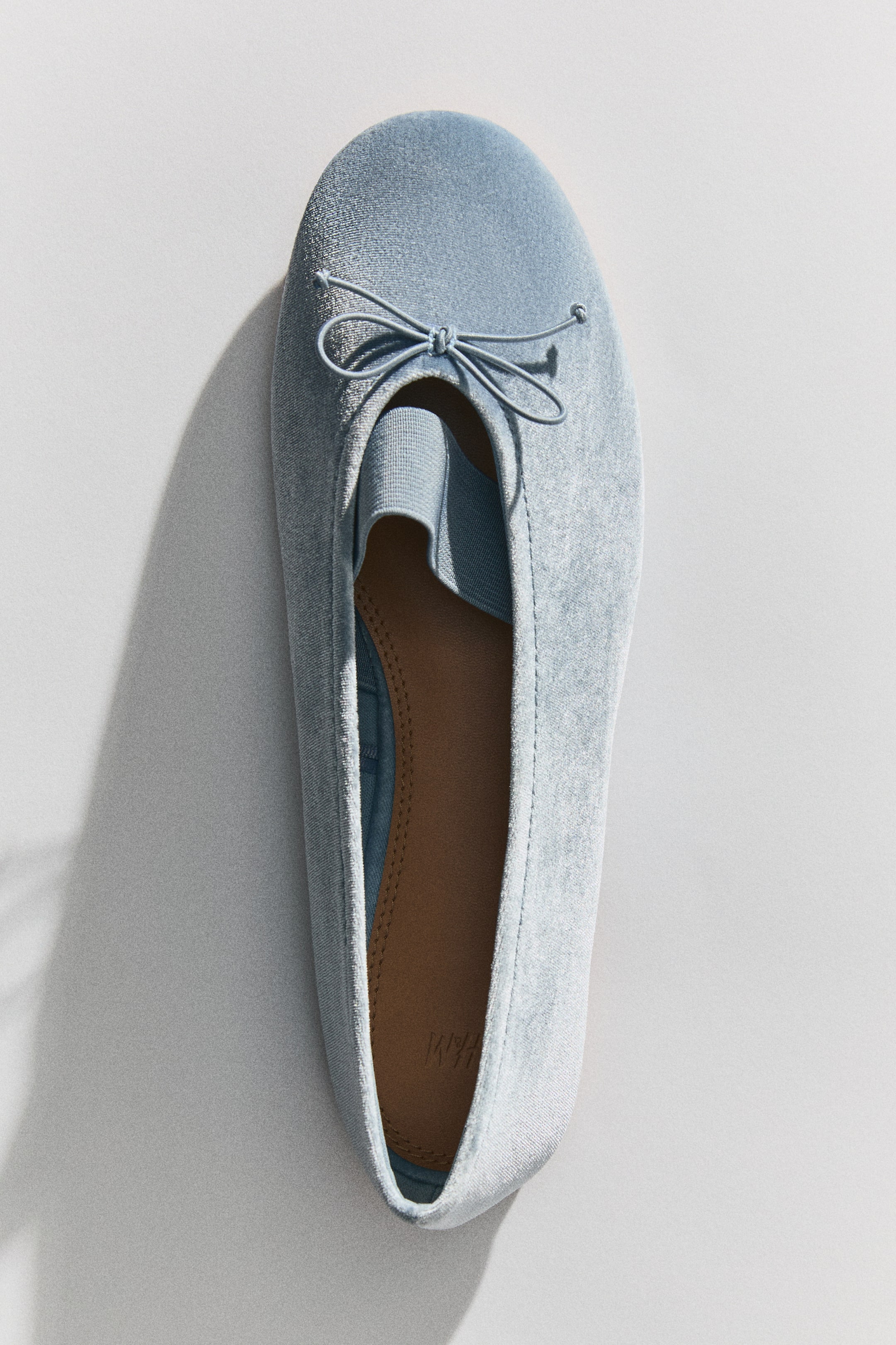 View larger image: A light blue ballet flat features soft velvet material, rounded toes, and a decorative bow at the front. A wide, elasticated foot strap is visible inside the opening.