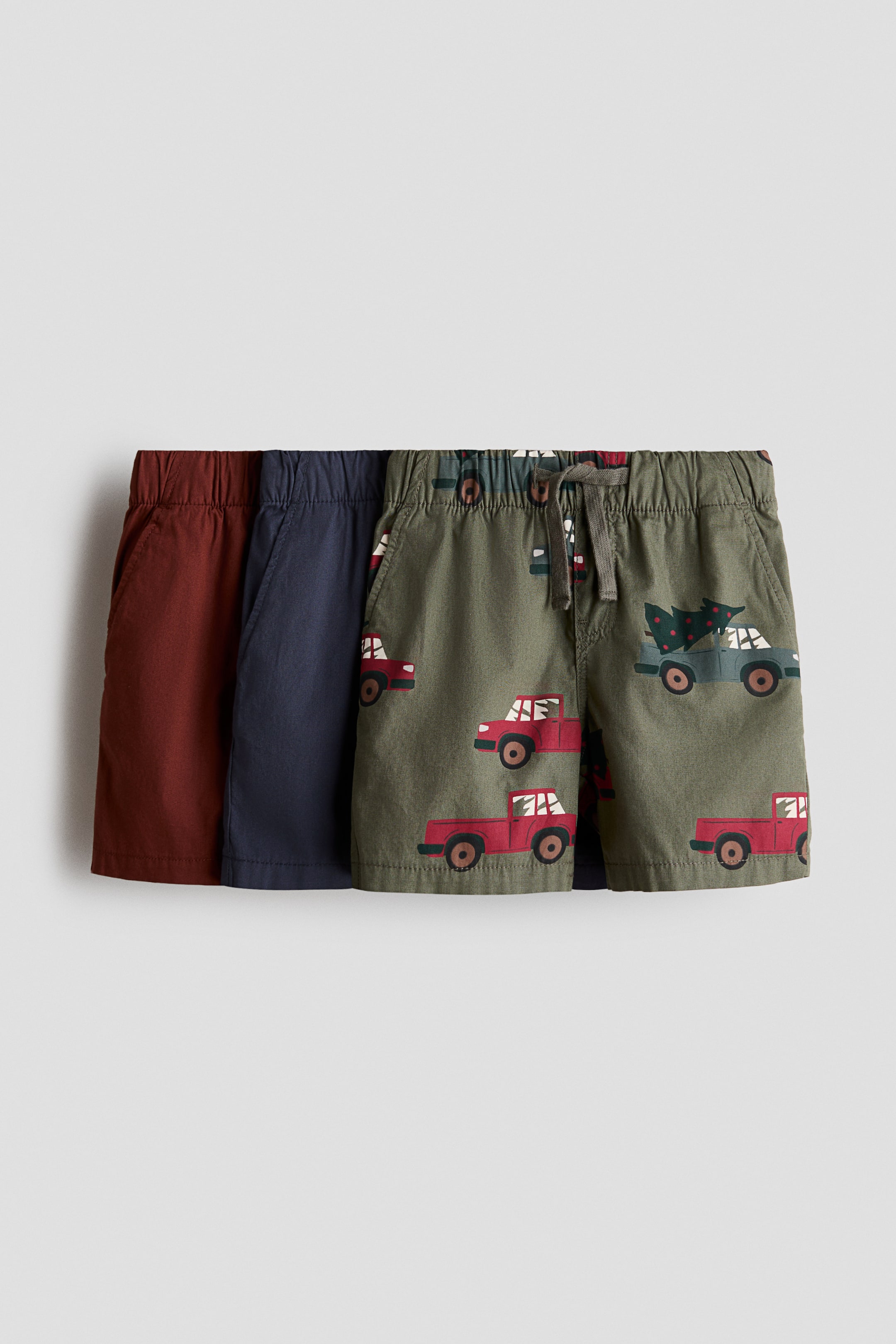 View larger image: Three pairs of cotton weave shorts are shown from the front, layered to display a solid rust-red pair, a solid navy blue pair, and an olive green pair featuring an all-over print of red and blue pickup trucks. The shorts are designed with an elasticated waistband, a drawstring on the front pair, a mock fly, and diagonal side pockets.