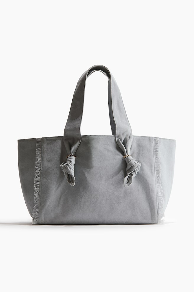 Un sac cabas gris clair est présenté de face, avec deux poignées supérieures, chacune nouée de manière décorative à travers un petit anneau de couleur laiton sur le devant du sac. Les côtés verticaux du sac présentent des coutures effilochées. - 1