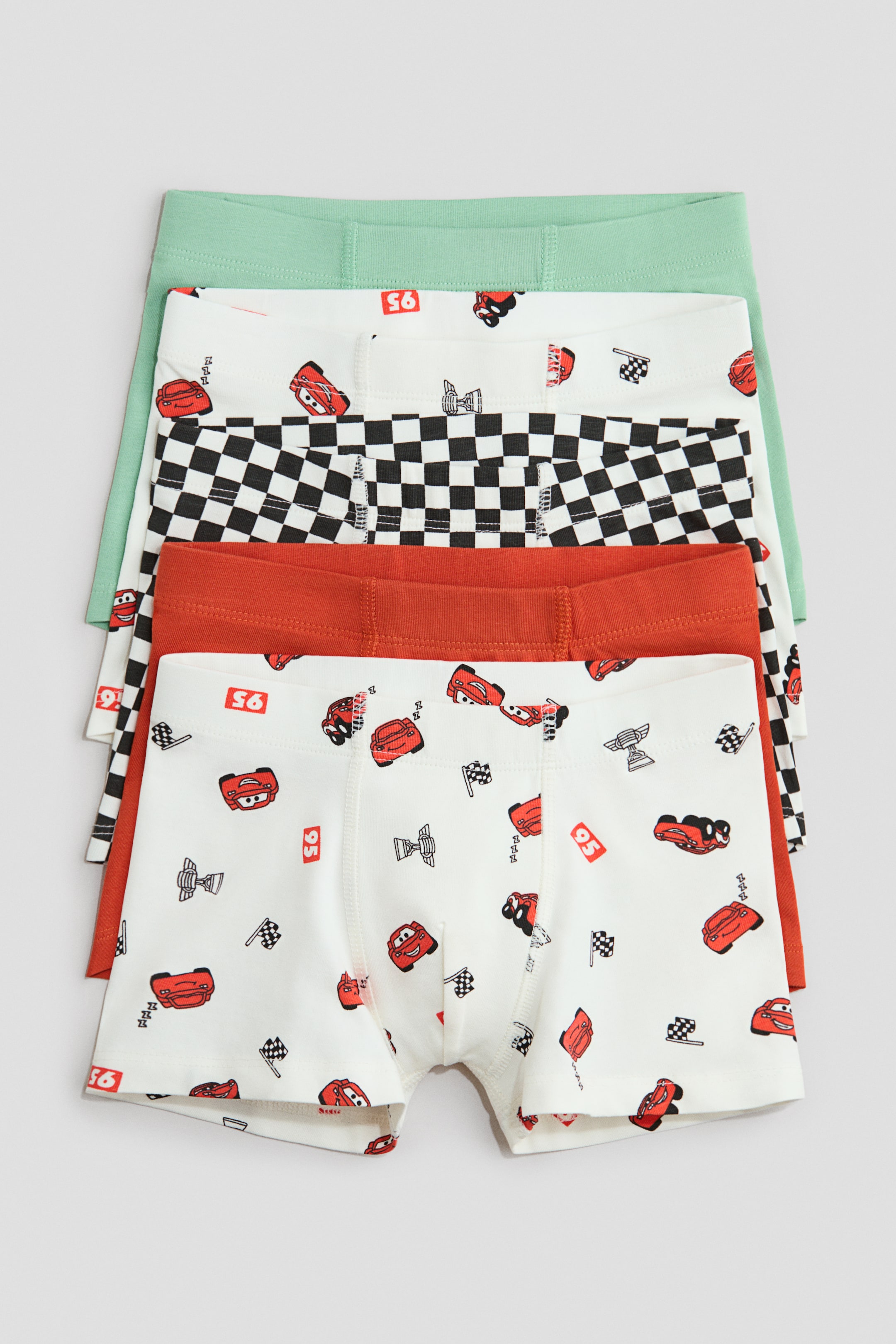 Xem ảnh lớn hơn: Một chồng bốn chiếc quần lót cotton jersey đang được trình bày, mỗi chiếc đều có phần thun che phủ ở eo, ống ngắn và mặt trước có lớp lót. Chiếc quần lót dưới cùng đang hướng MẶT TRƯỚC, hiển thị nền trắng với họa tiết xe hơi hoạt hình màu đỏ, trong khi các cặp quần lót nhìn thấy một phần phía trên có thiết kế màu xanh nhạt trơn, kẻ ô đen trắng, và màu cam gỉ trơn.