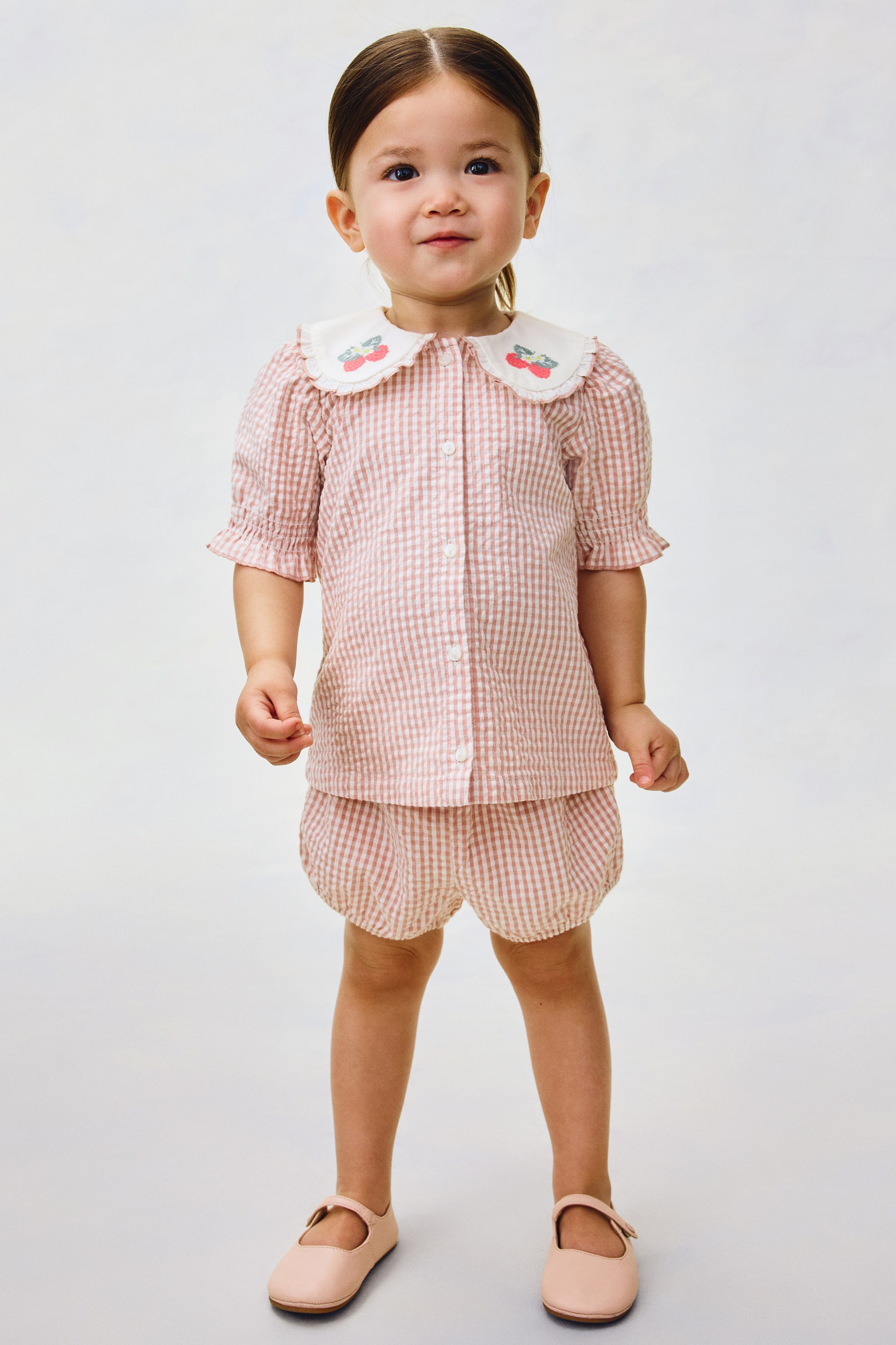 View larger image: The two-piece children's outfit features a pink and white gingham check. The shirt is designed with a white rounded collar showcasing embroidered cherries, puffed short sleeves, and ruffled cuffs, while the matching bloomer shorts display elasticated leg openings.