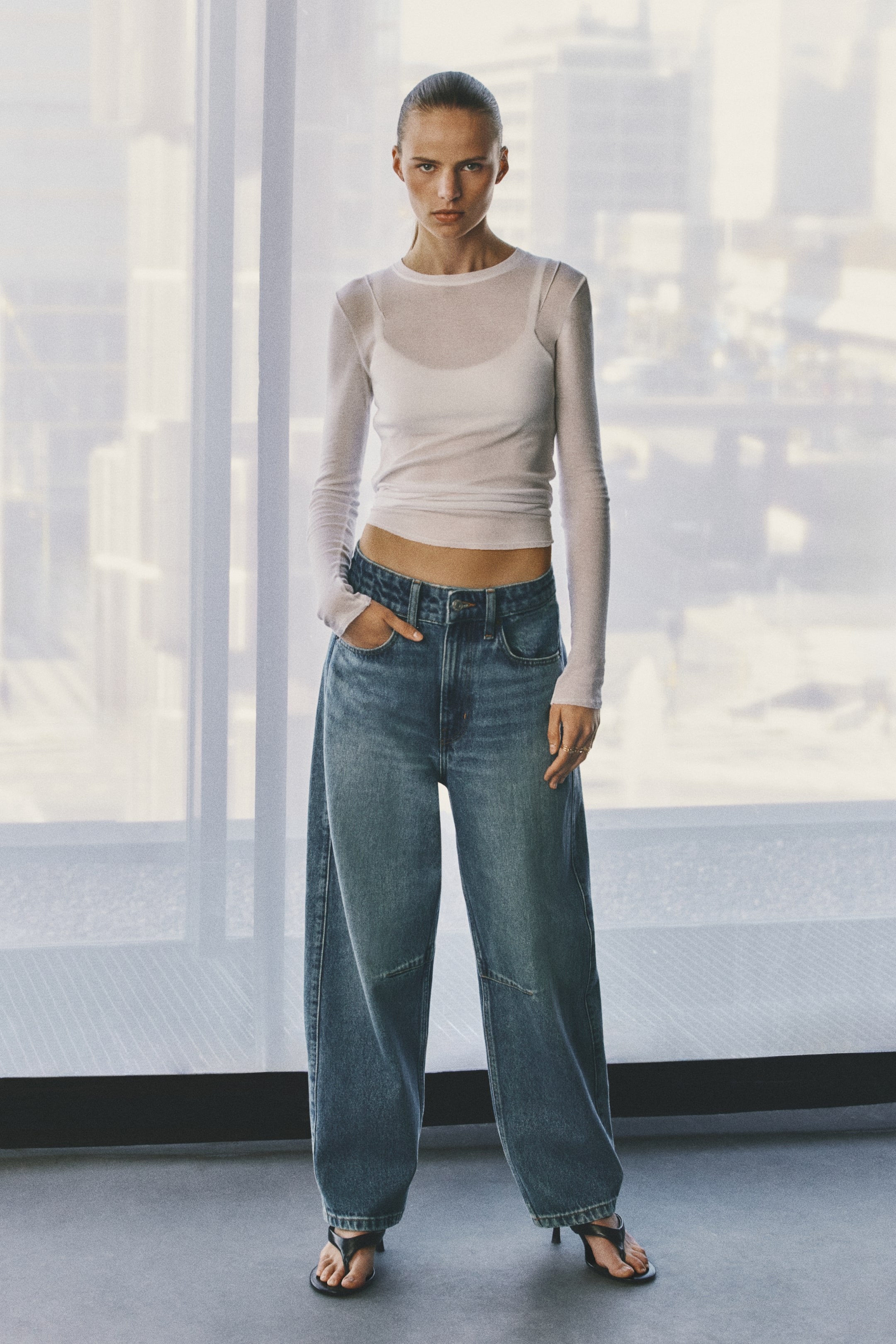 Voir une image plus grande: Jean en denim bleu taille haute à coupe ample, avec des jambes volumineuses et arrondies qui se resserrent à l’ourlet, porté par le mannequin. Le mannequin est debout, une main appuyée dans une poche avant, et agencé le jean avec un haut blanc semi-transparent à manches longues et des sandales noires à talons.