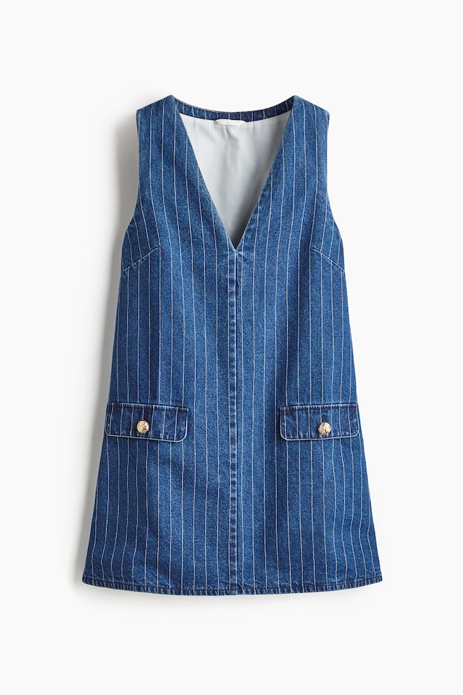 Een blauwe denim tuinjurk toont witte krijtstrepen aan de voorkant en heeft een diepe V-hals en twee opgestikte zakken, elk versierd met een goudkleurige knoop. - 1
