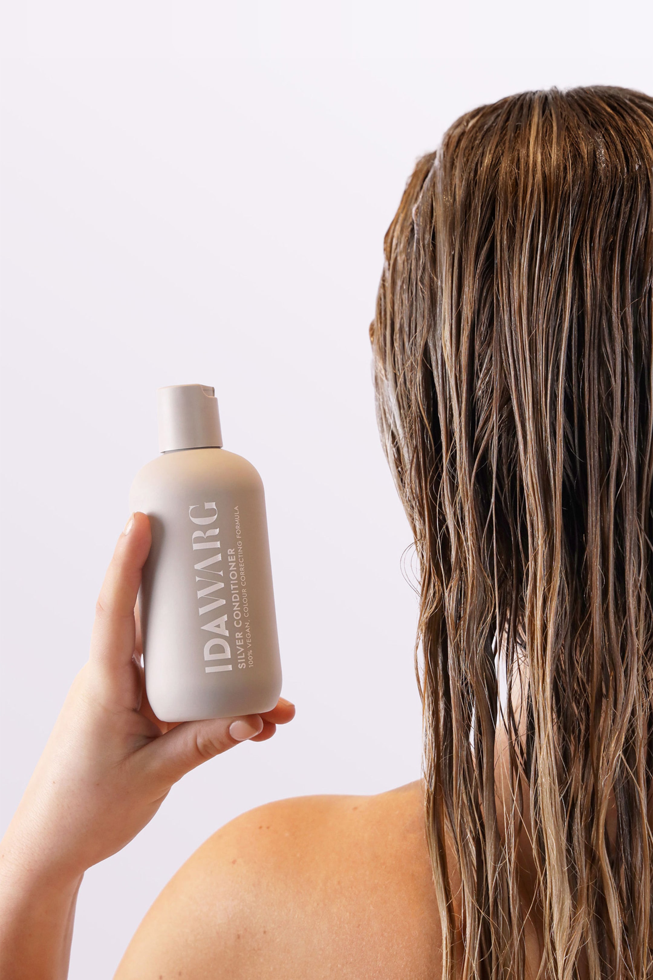 Größeres Bild ansehen: Eine hellgraue, abgerundete rechteckige Flasche IDAW�RG Silver Conditioner wird von einer Hand gehalten, die ihr Frontetikett präsentiert. Die Flasche zeigt einen Klappverschluss und "IDAW�RG SILVER CONDITIONER 100% VEGAN. COLOUR CORRECTING FORMULA" in dunkelgrauer Schrift.