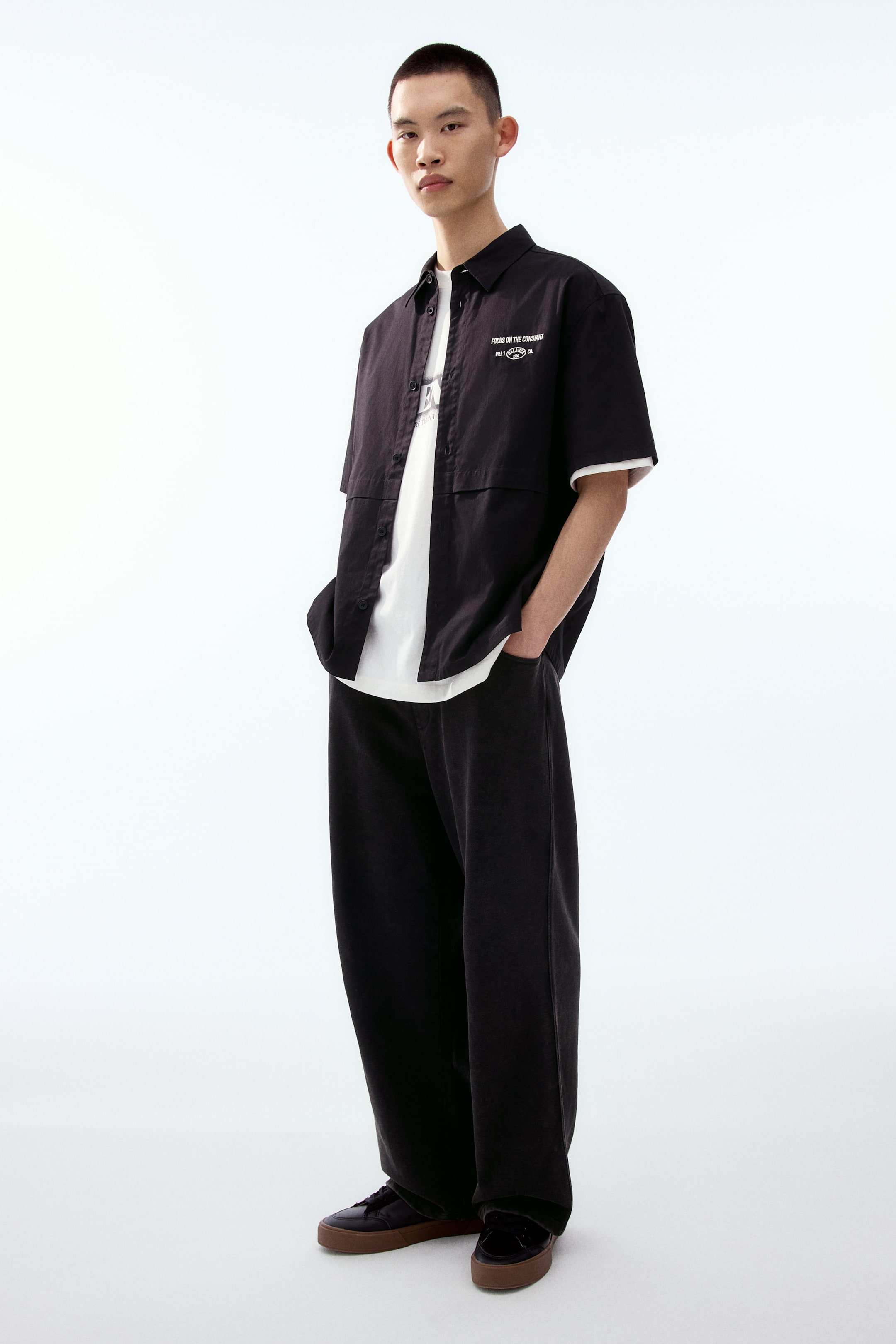 拡大画像を表示: モデルはターンダウンカラー、クラシックな前開きボタン、前身頃の水平プリーツ、左胸の刺繍モチーフが施された黒の半袖シャツを着用しています。シャツは白いTシャツの上に羽織るように前を開けて着ており、裾はストレートカットでゆったりとしたフィットです。