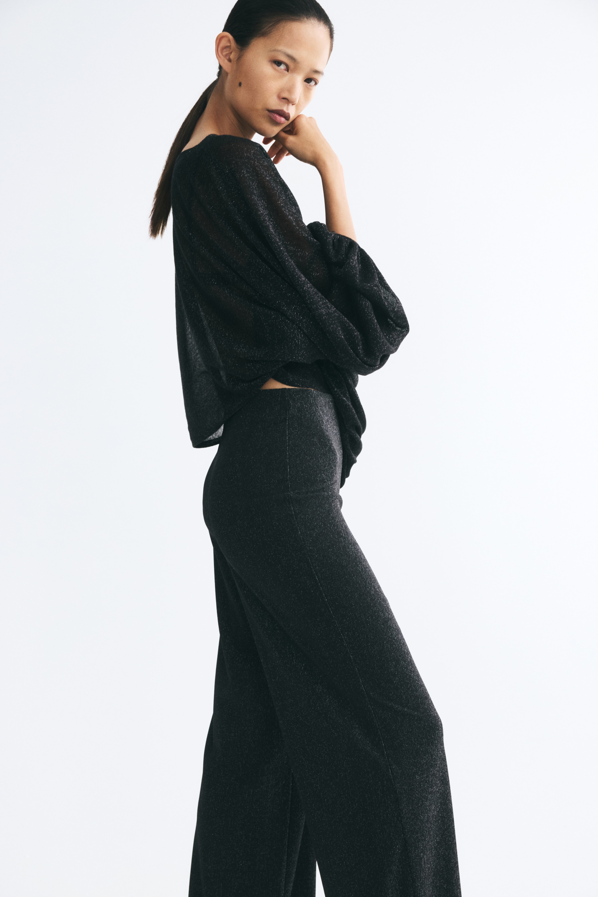 Loose-fit trousers made from a shimmering black fabric are hanging in a front view, showing their wide-leg silhouette. The trousers feature a clean, elasticated waistband and a relaxed, straight cut.