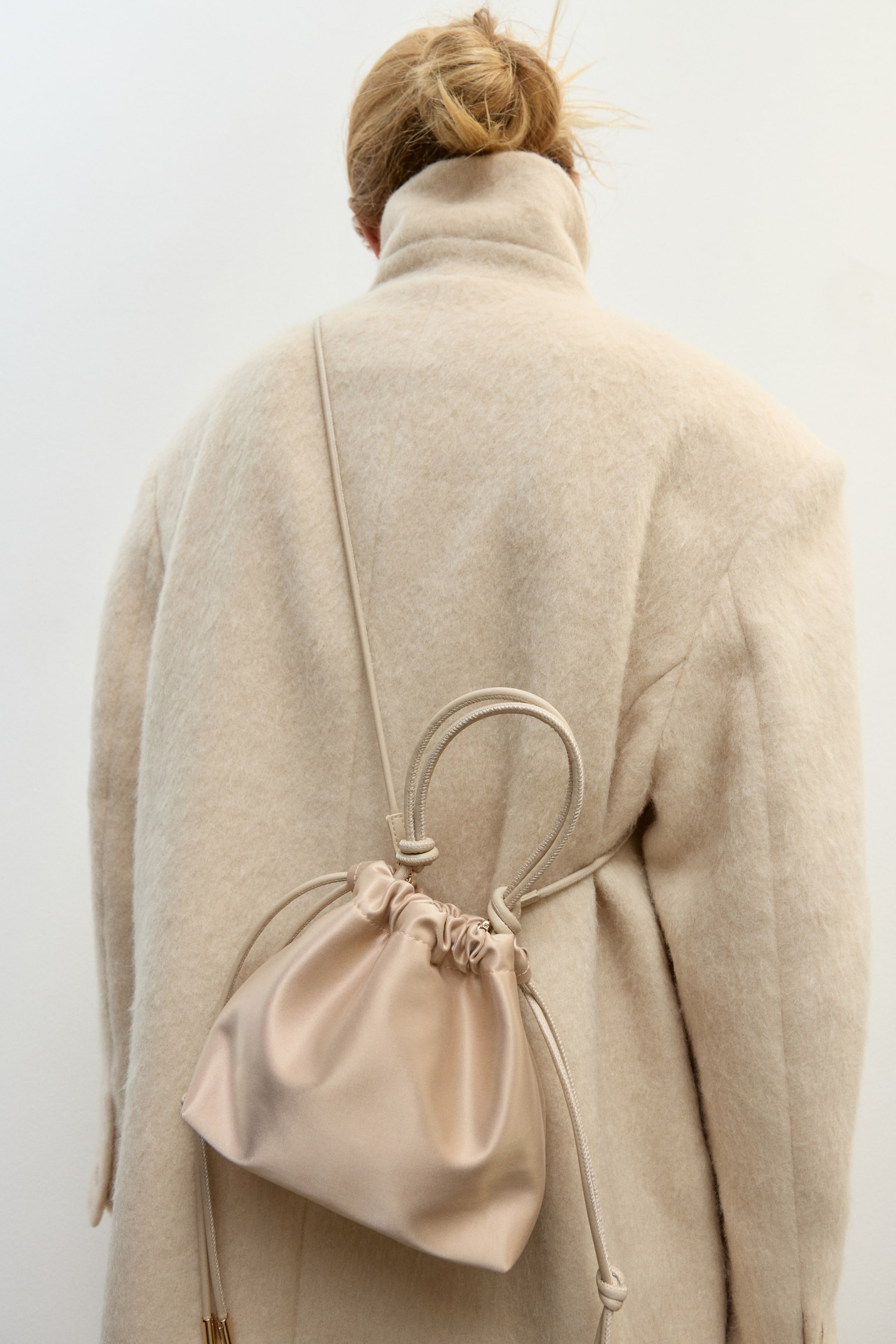 Se større billede: En lille, lysebeige satin crossbodytaske bæres af modellen over en tekstureret cremefarvet frakke. Tasken har en rynket snørelukning, en tynd skulderrem, der er draperet over skulderen, og et løst tophåndtag.