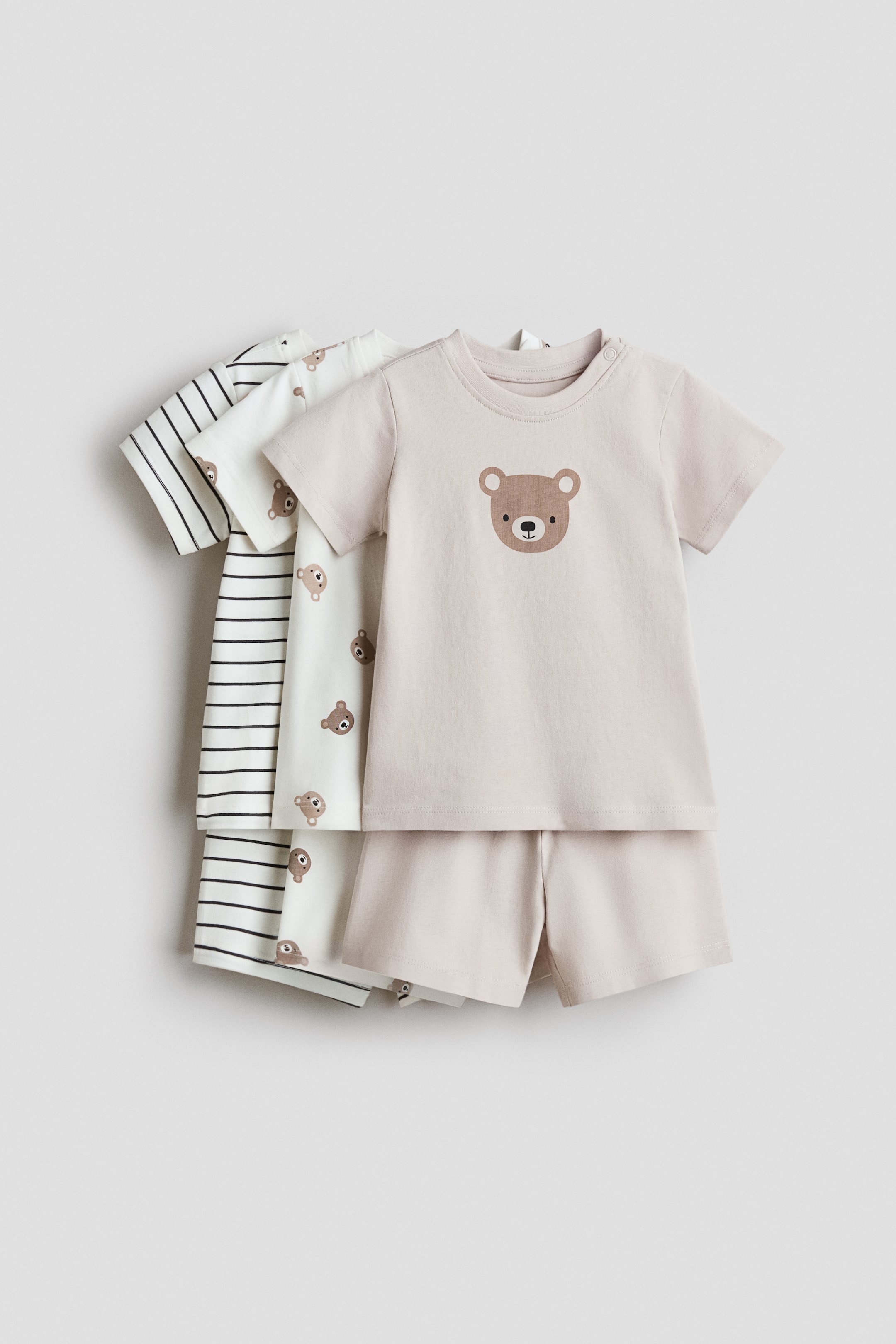 拡大画像を表示: 3セットのベビー服が展示されており、前列のセットはライトベージュの半袖トップスに茶色のクマの顔のグラフィックがフロントに施され、マッチングショーツが付いています。その後ろには、2枚の追加の白い半袖トップスが重なっており、一つは小さなクマの顔がパターン化され、もう一つは黒の水平ストライプと小さなクマの顔のグラフィックが施されています。