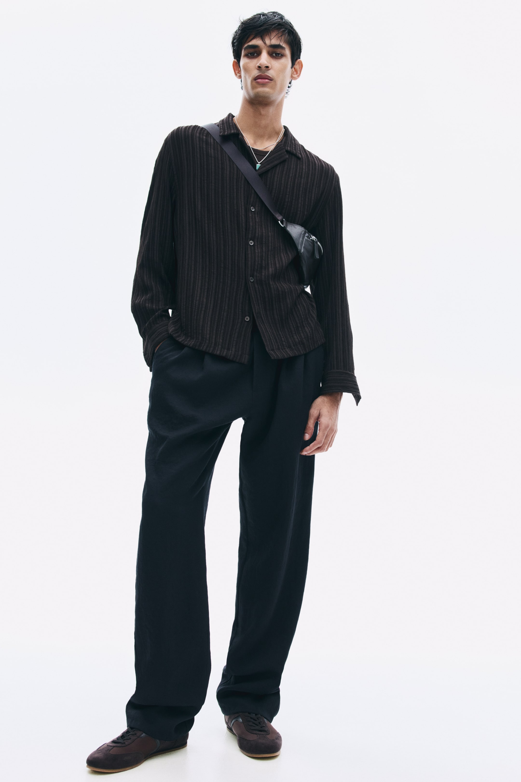 View larger image: The model is wearing dark relaxed-fit pants in a viscose blend twill, standing with one hand placed in a diagonal side pocket. The pants feature a straight-leg silhouette and are styled with a dark, textured long-sleeve button-up shirt and a small crossbody bag.