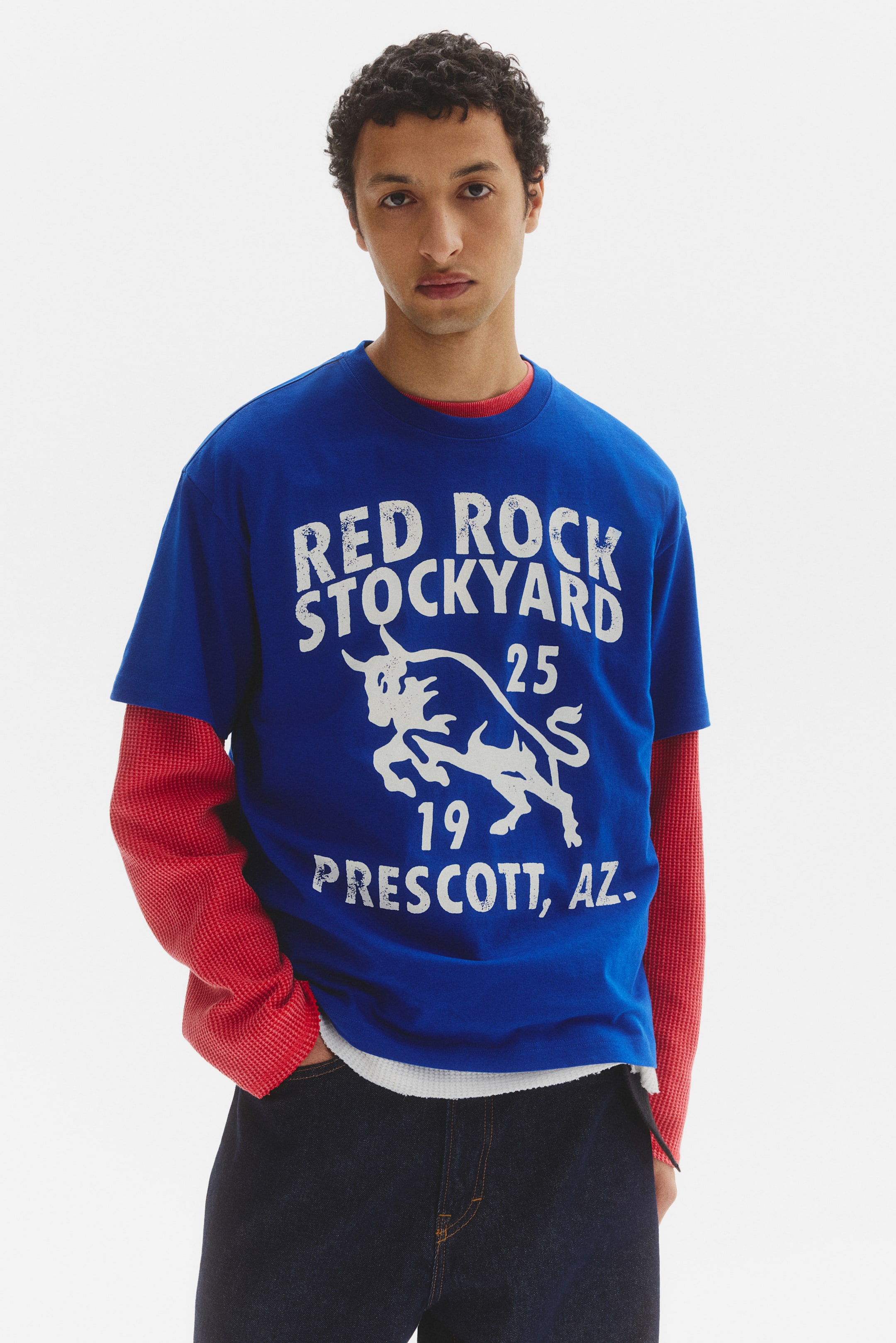 View larger image: A blue crew-neck t-shirt showcases a white distressed graphic on its front, featuring a bull design with the text "RED ROCK STOCKYARD 25 19 PRESCOTT, AZ." It is layered over a red waffle-knit long-sleeve shirt, with the red sleeves and collar peeking through.