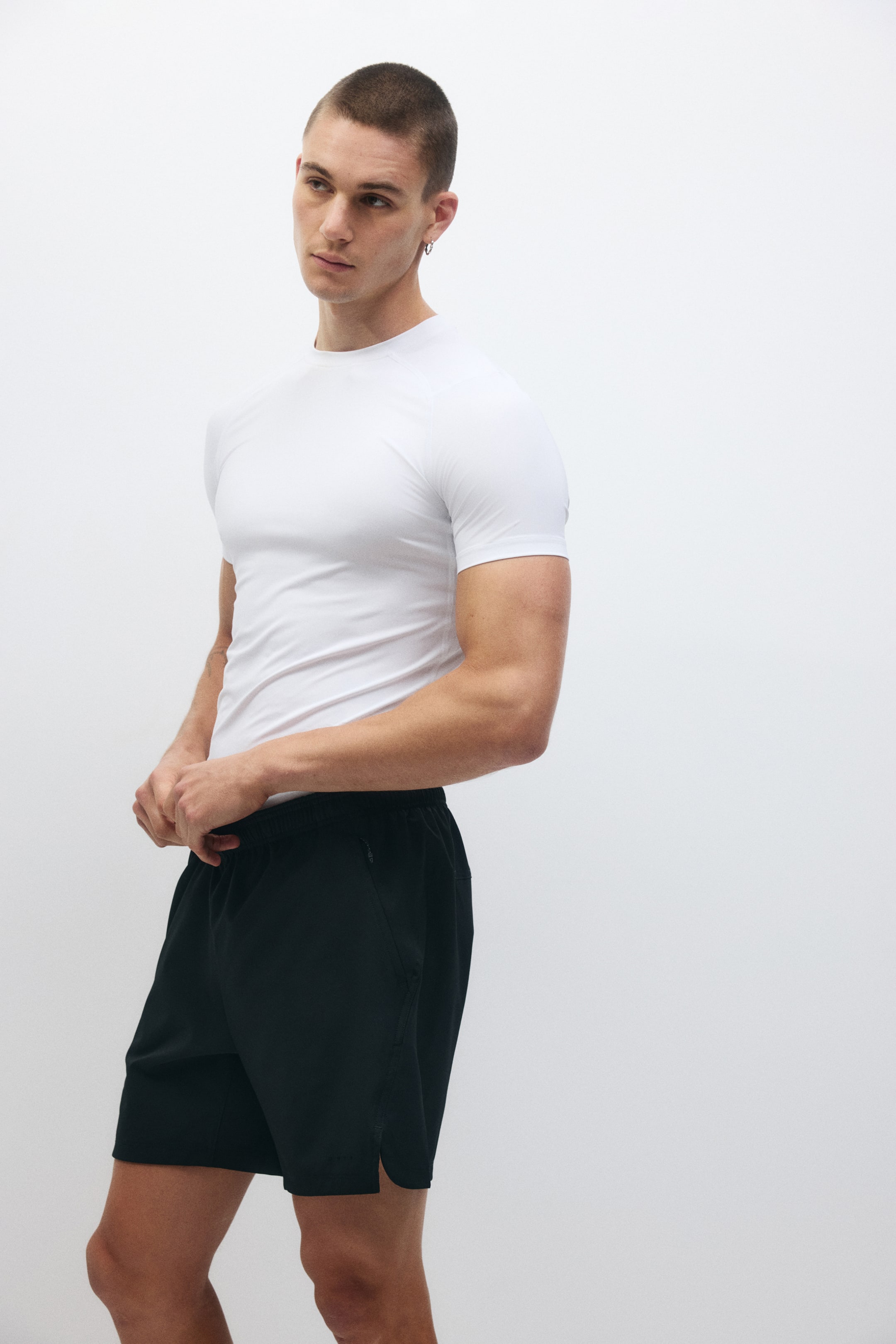 View larger image: A snug-fitting white crew neck tee with short sleeves adorns the upper body. Complementing it are black athletic shorts, sitting comfortably at the waist. These shorts boast an elastic waistband, a zippered side pocket, and subtle side slits at the hem, ensuring both style and functionality.