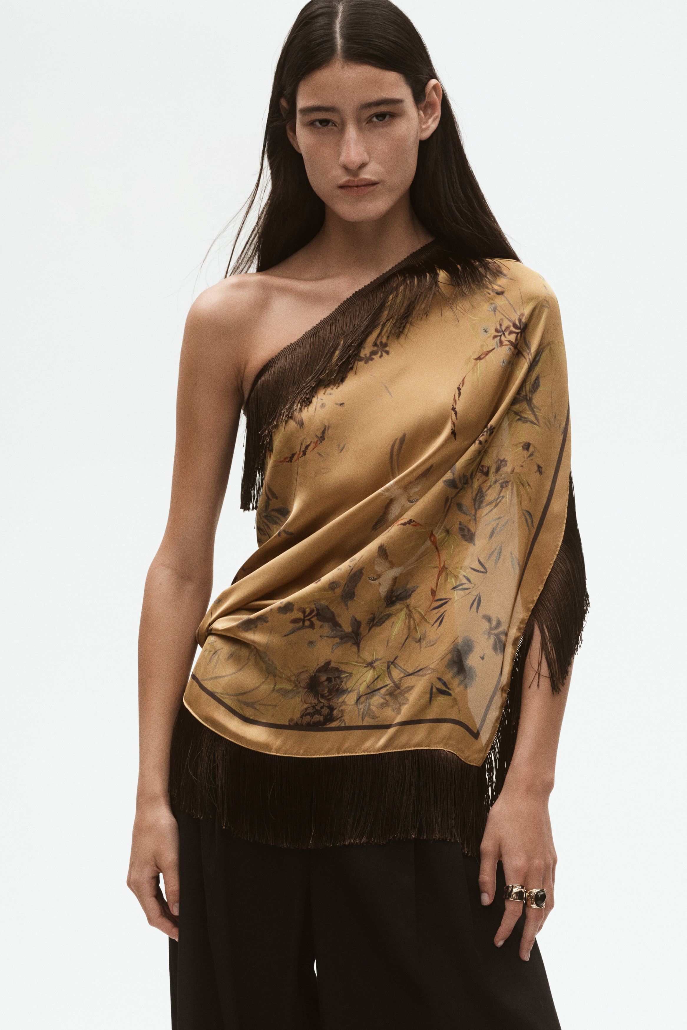 A golden-brown silk scarf featuring a subtle botanical print is displayed from the front, loosely knotted at the top to form a draped triangular shape. Long, dark brown fringe borders the edges, cascading downward and outward.