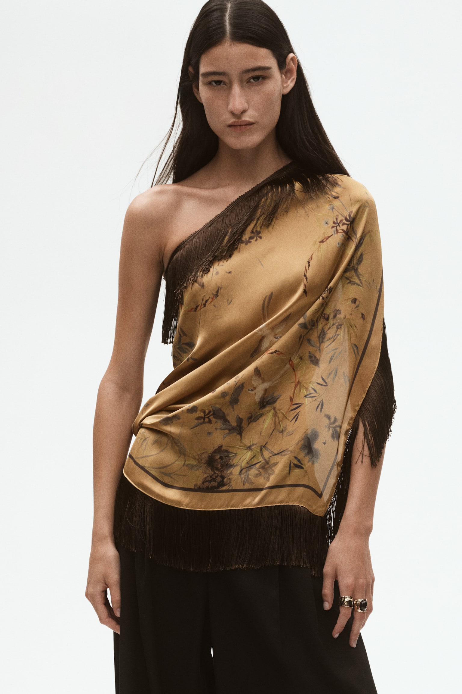 A golden-brown silk scarf with a subtle botanical print is shown from the front, loosely knotted at the top to form a draped triangular shape. Long, dark brown fringe borders the edges, cascading downward and outward. - 1
