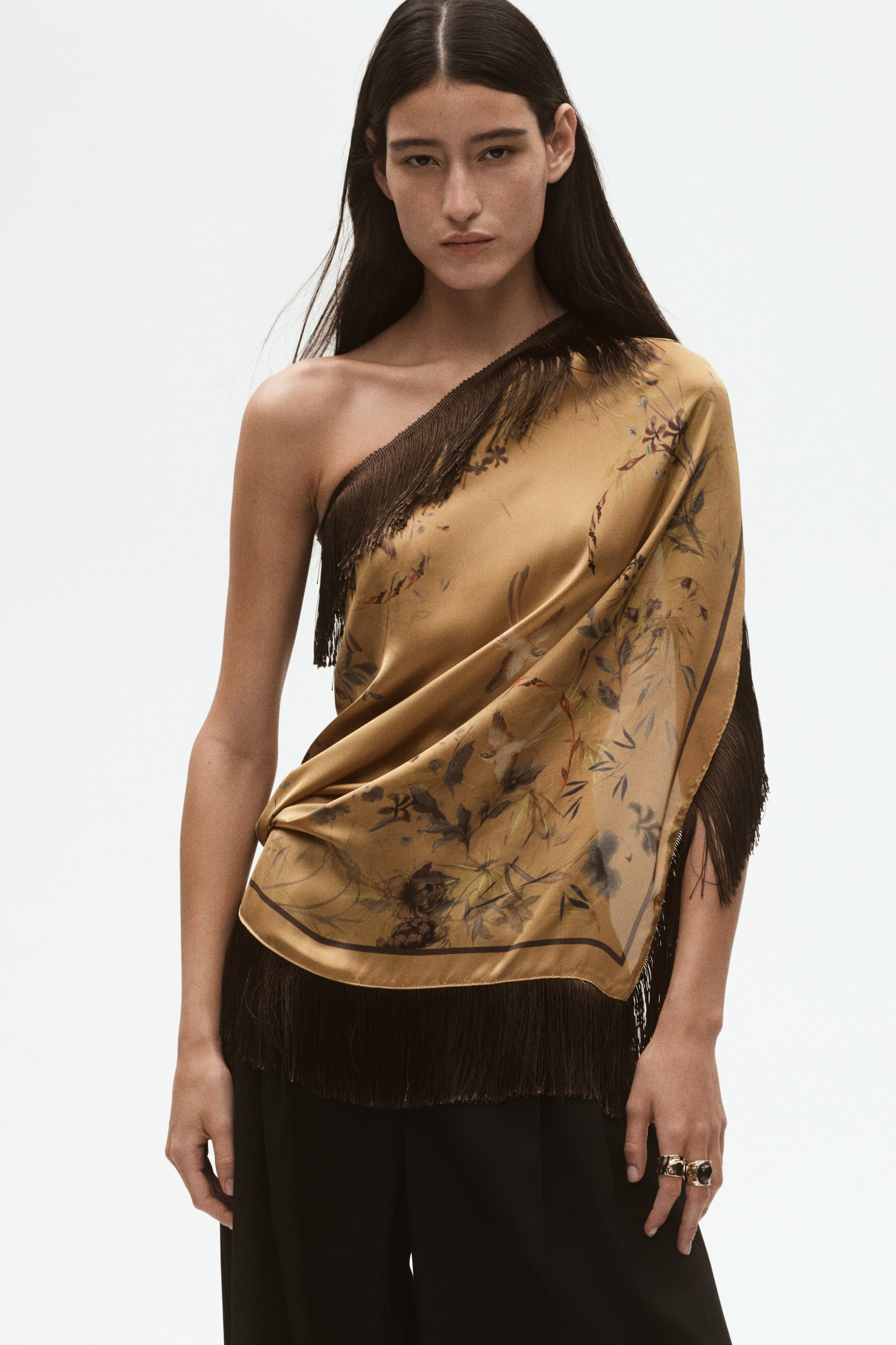 View larger image: A golden-brown satin square scarf with a botanical print and dark brown fringe is styled on the model as a draped, one-shoulder top. The scarf is wrapped diagonally across the torso and paired with black trousers.