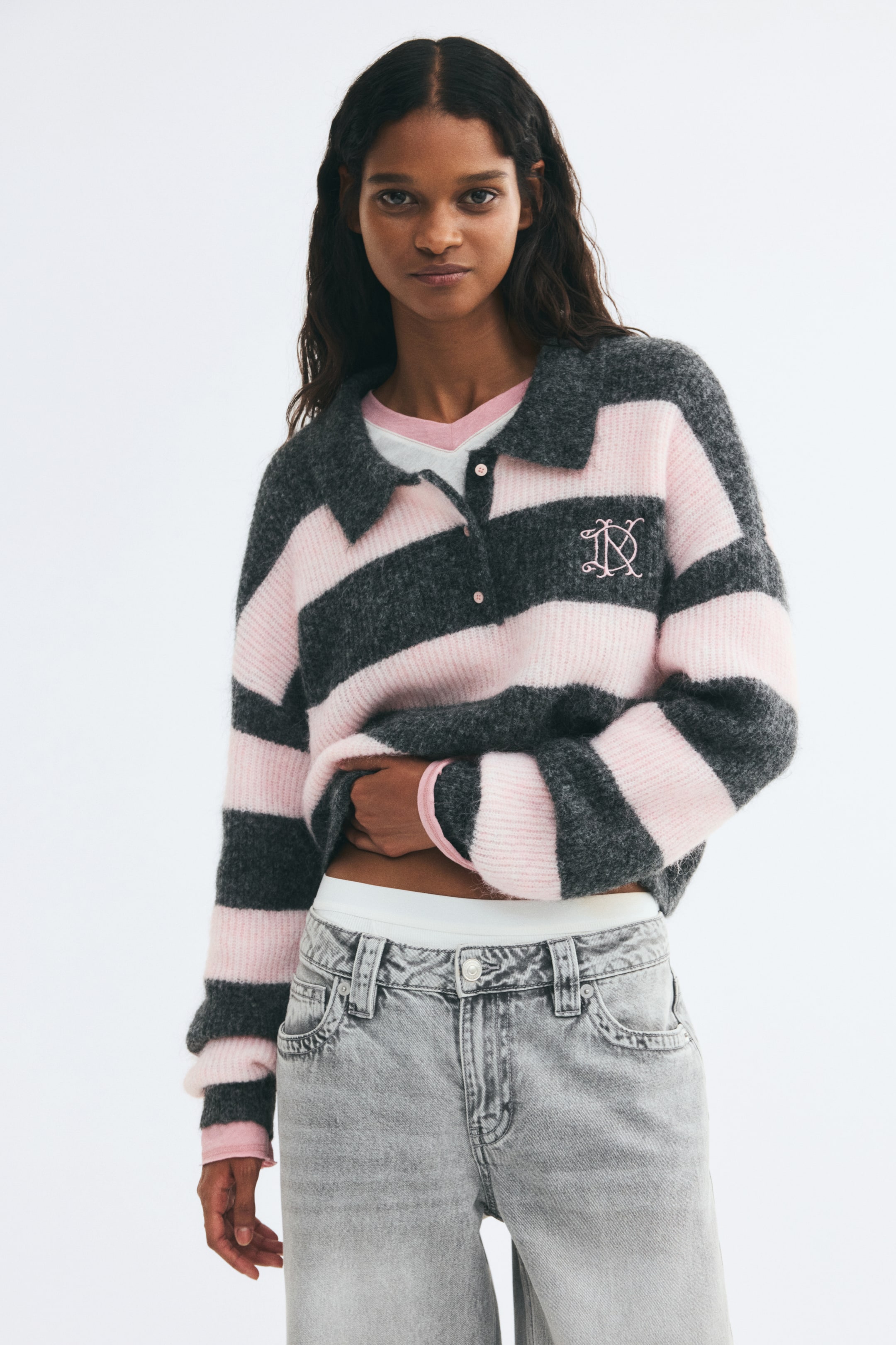 Agrandir l'image: Un pull polo à coupe décontractée en maille côtelée rayée gris et rose est porté par le mannequin, qui tient légèrement l’ourlet avant relevé. Le pull présente un col, une patte de boutonnage, des manches longues et un monogramme rose brodé sur la poitrine, porté sur un t-shirt blanc et rose et un jean gris.