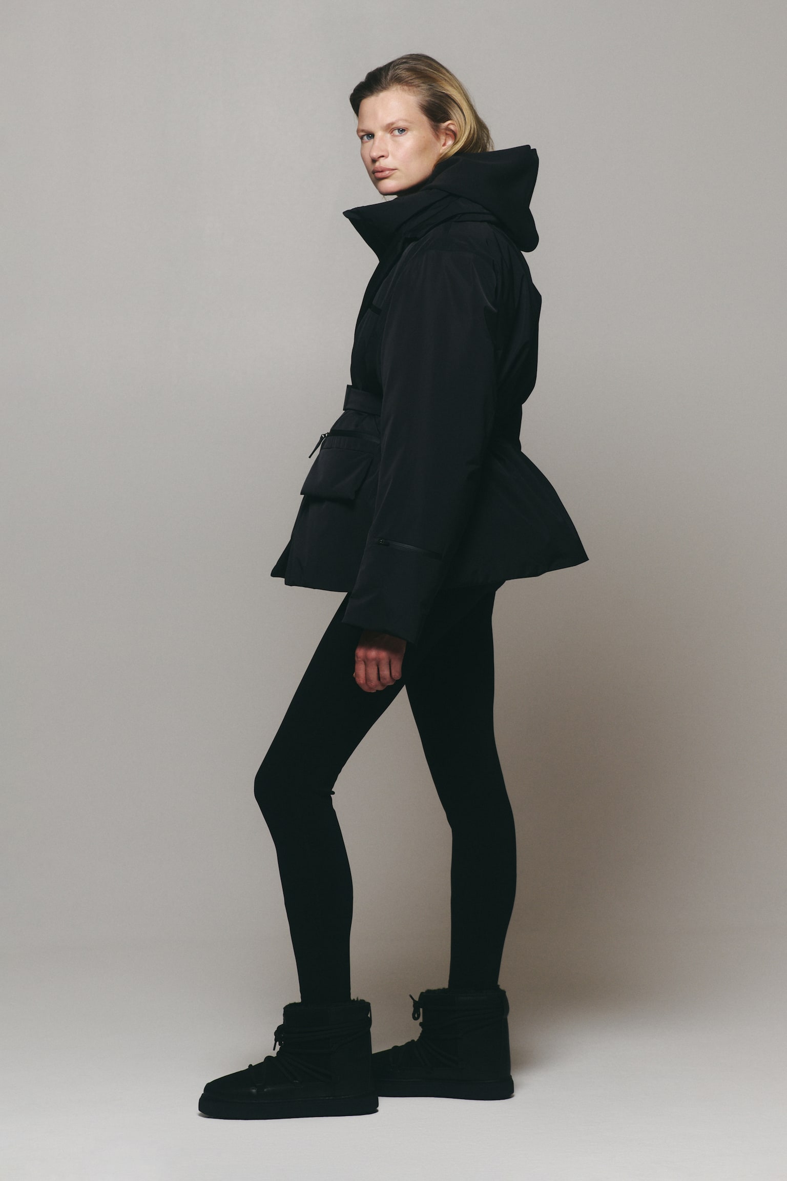 Une veste dâhiver noire à capuche, dotée dâun col montant, dâune taille ceinturée et dâun ourlet évasé, est portée par le mannequin. La veste est associée à des leggings noirs ajustés et à des bottes dâhiver noires à lacets. - 1