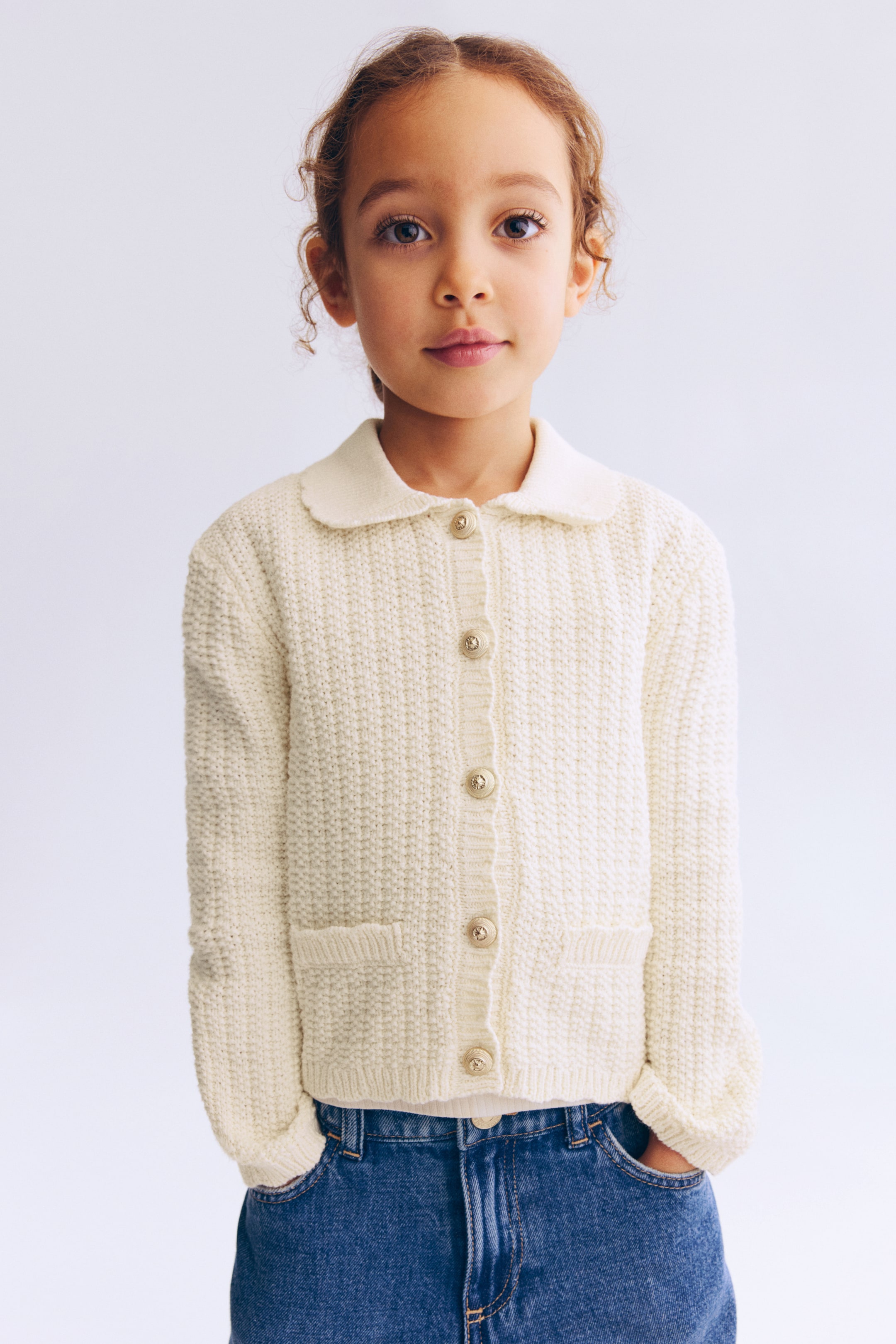 Ampliar la imagen: Un cárdigan color crema muestra su FRENTE, con un tejido suave y texturizado, un cuello acanalado con borde festoneado, botones metálicos con relieve que recorren la parte frontal, bolsillos frontales decorativos con vivos acanalados, puños estrechos acanalados y un dobladillo acanalado.