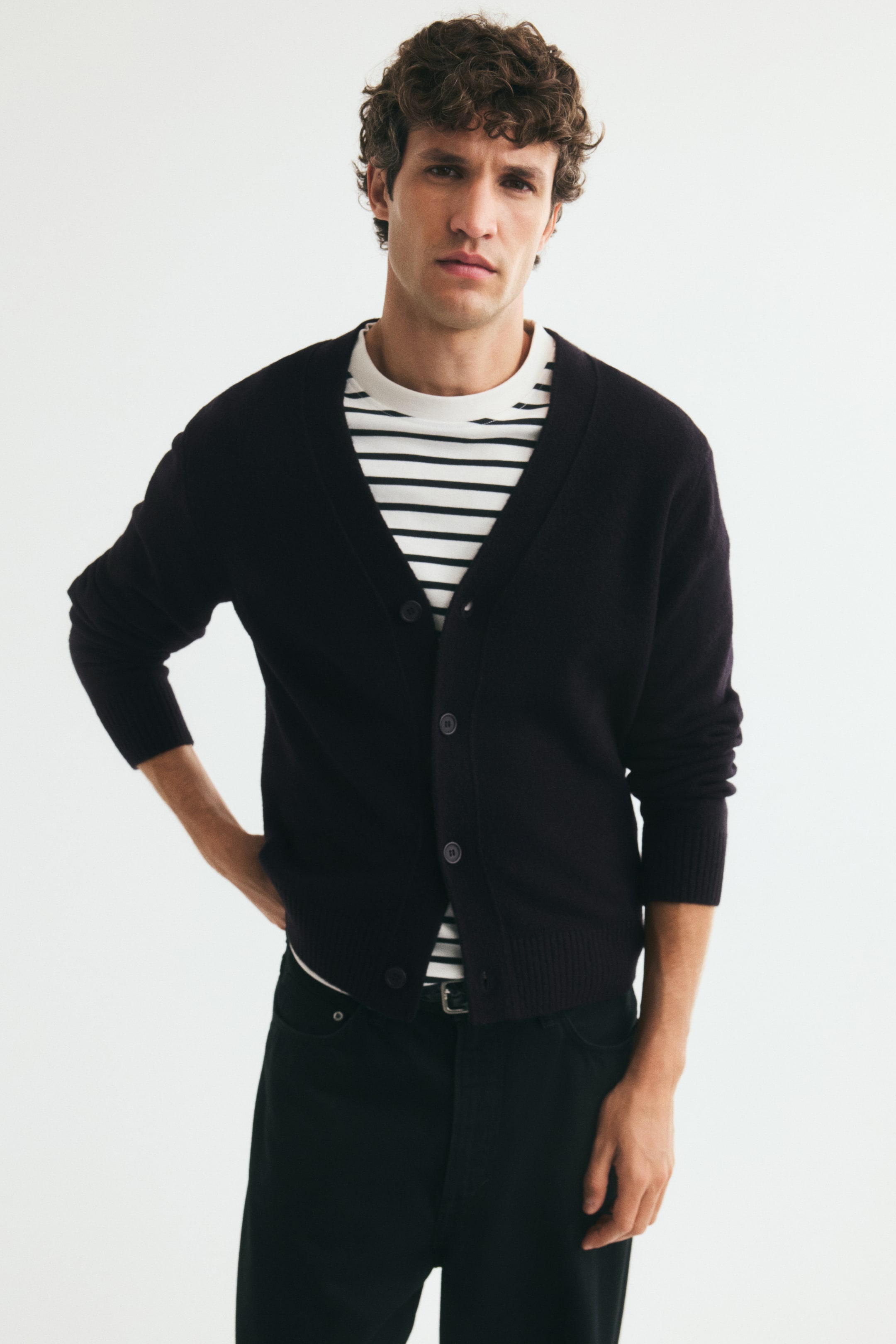 Größeres Bild anzeigen: Ein dunkelvioletter Strick-Cardigan mit V-Ausschnitt und durchgehender Knopfleiste wird von dem Model über einem schwarz-weiß gestreiften T-Shirt getragen. Der Cardigan ist teilweise zugeknöpft gestylt und zeigt seine reguläre Passform, gerippten Bündchen und den gerippten Saum.