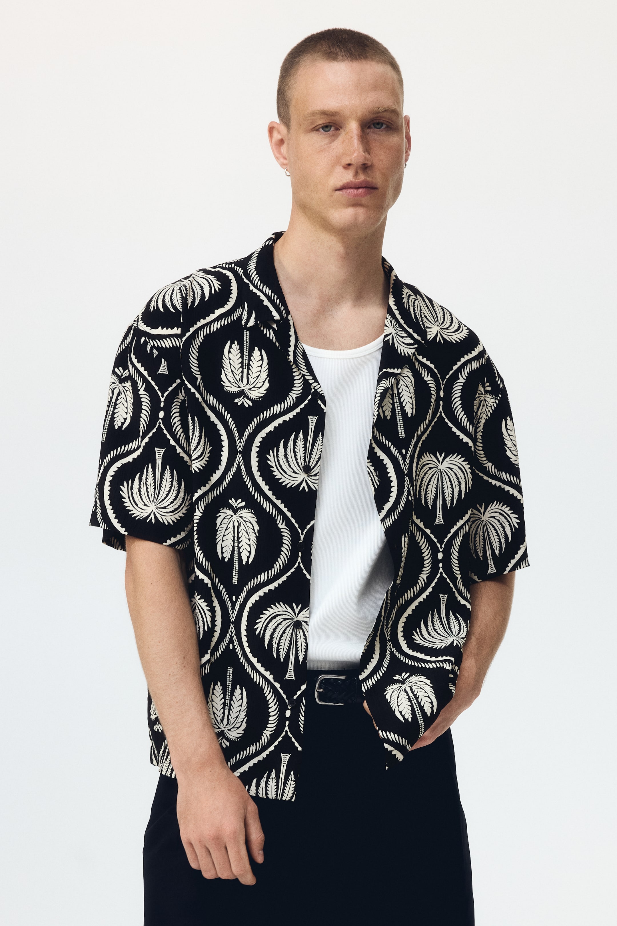 View larger image: A black short-sleeve shirt is shown facing forward, featuring a white palm tree and decorative scrollwork pattern. It is styled with a camp collar and is worn open over a white undershirt.