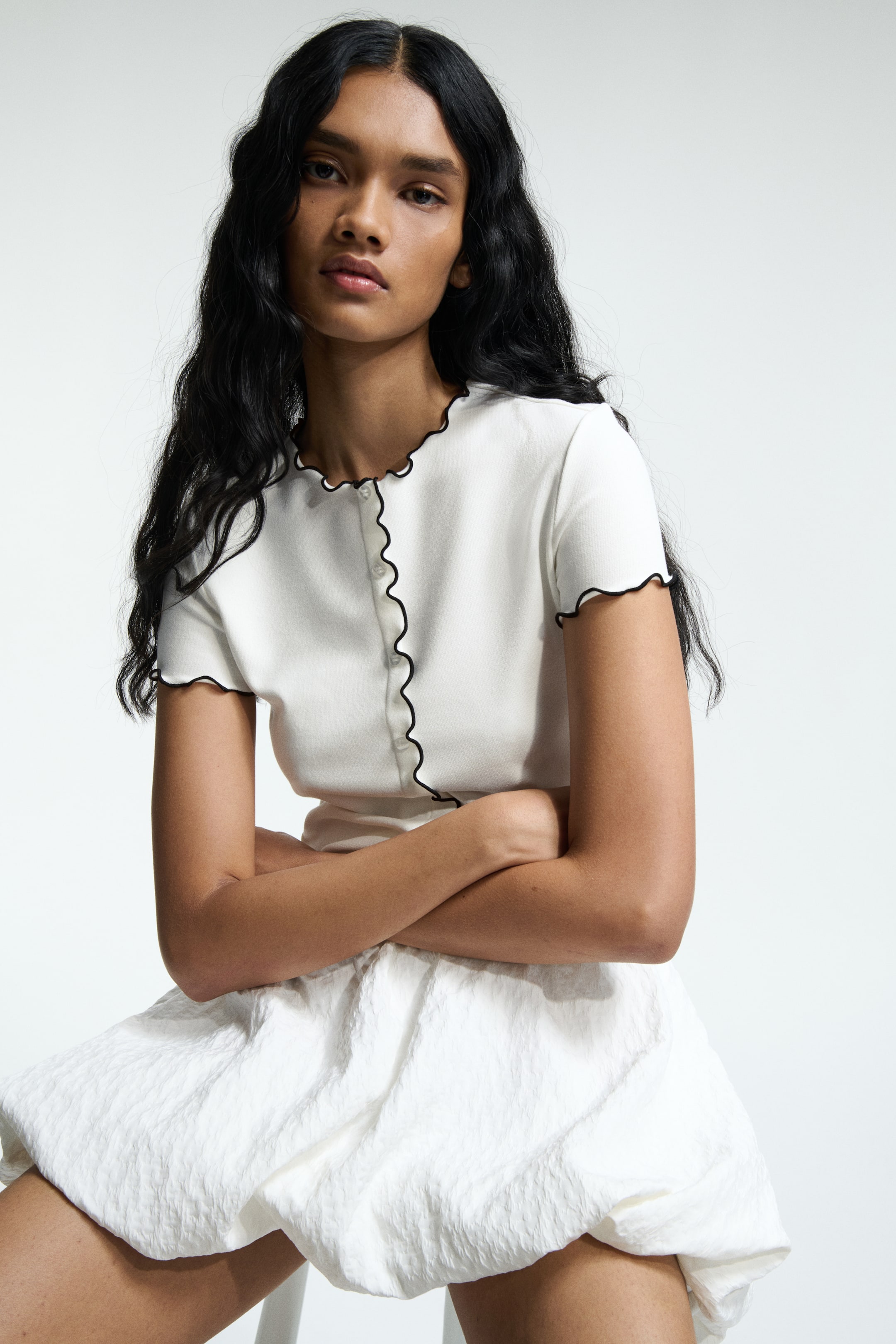 View larger image: A white short-sleeved cardigan is featuring a round neck and button-front closure, detailed with black wavy trim along the neckline, placket, and sleeve hems. It is worn with a white voluminous mini skirt, showcasing a textured, crinkled fabric.