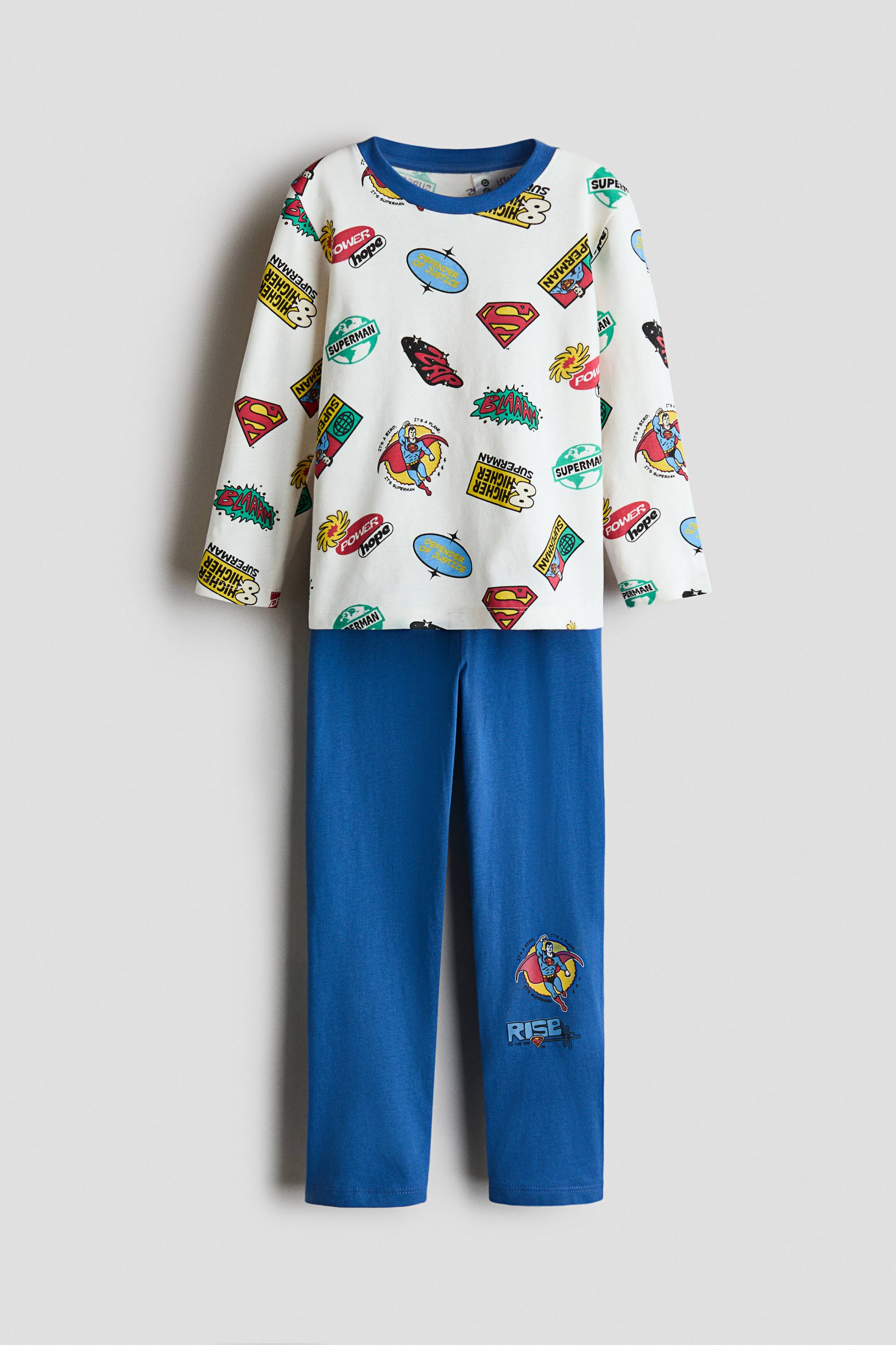 View larger image: A two-piece pajama set featuring a white long-sleeve top with a blue ribbed crew neck and an all-over print of various Superman logos and graphics. The coordinating blue pajama pants are straight-legged and feature a small Superman graphic with the text “RISE” on the lower right leg.