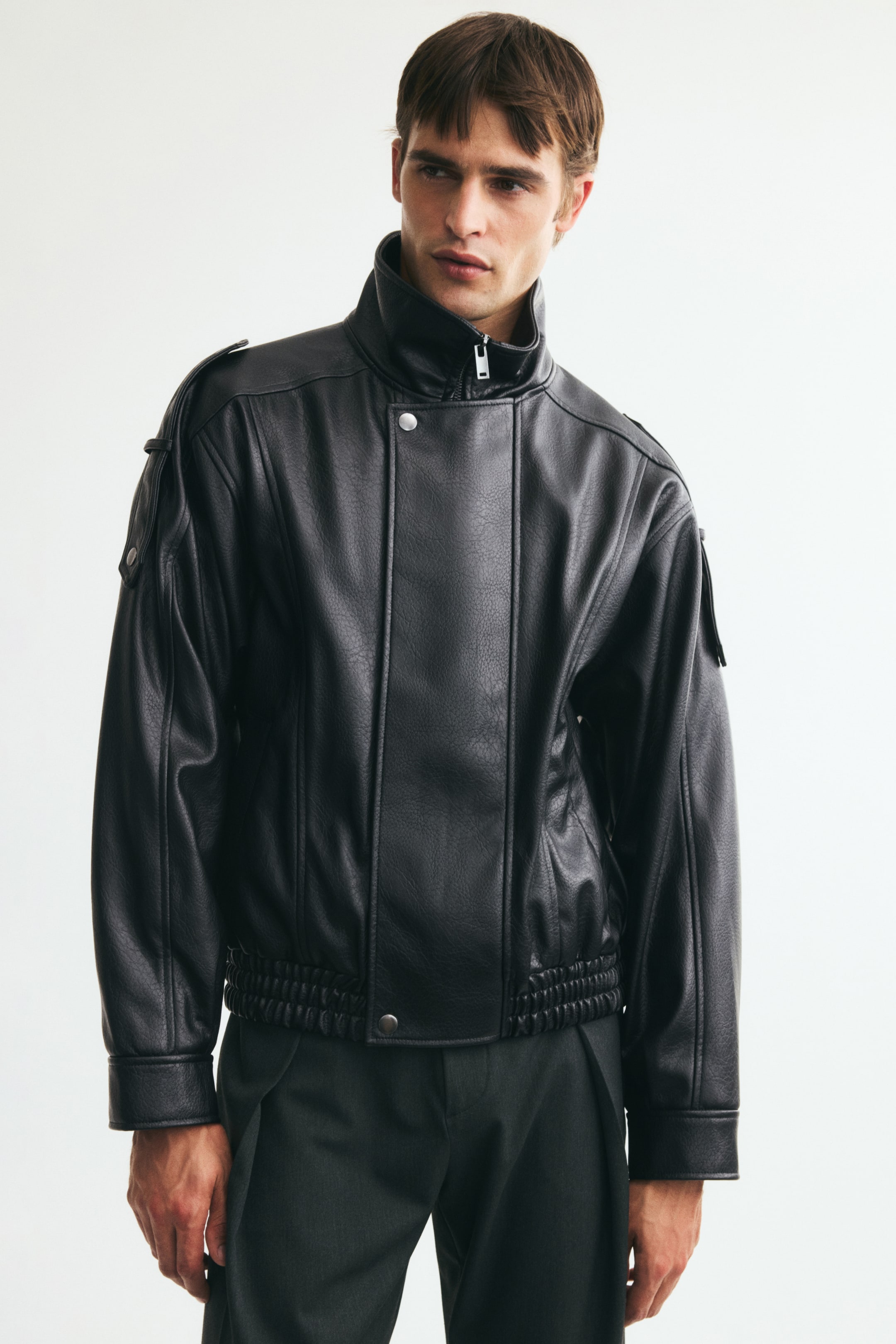 View larger image: The model is wearing a black jacket in coated fabric, featuring visible seams, a stand collar, a partially zipped front with press-studs, epaulettes with press-studs, and smocked elastication at the hem.
