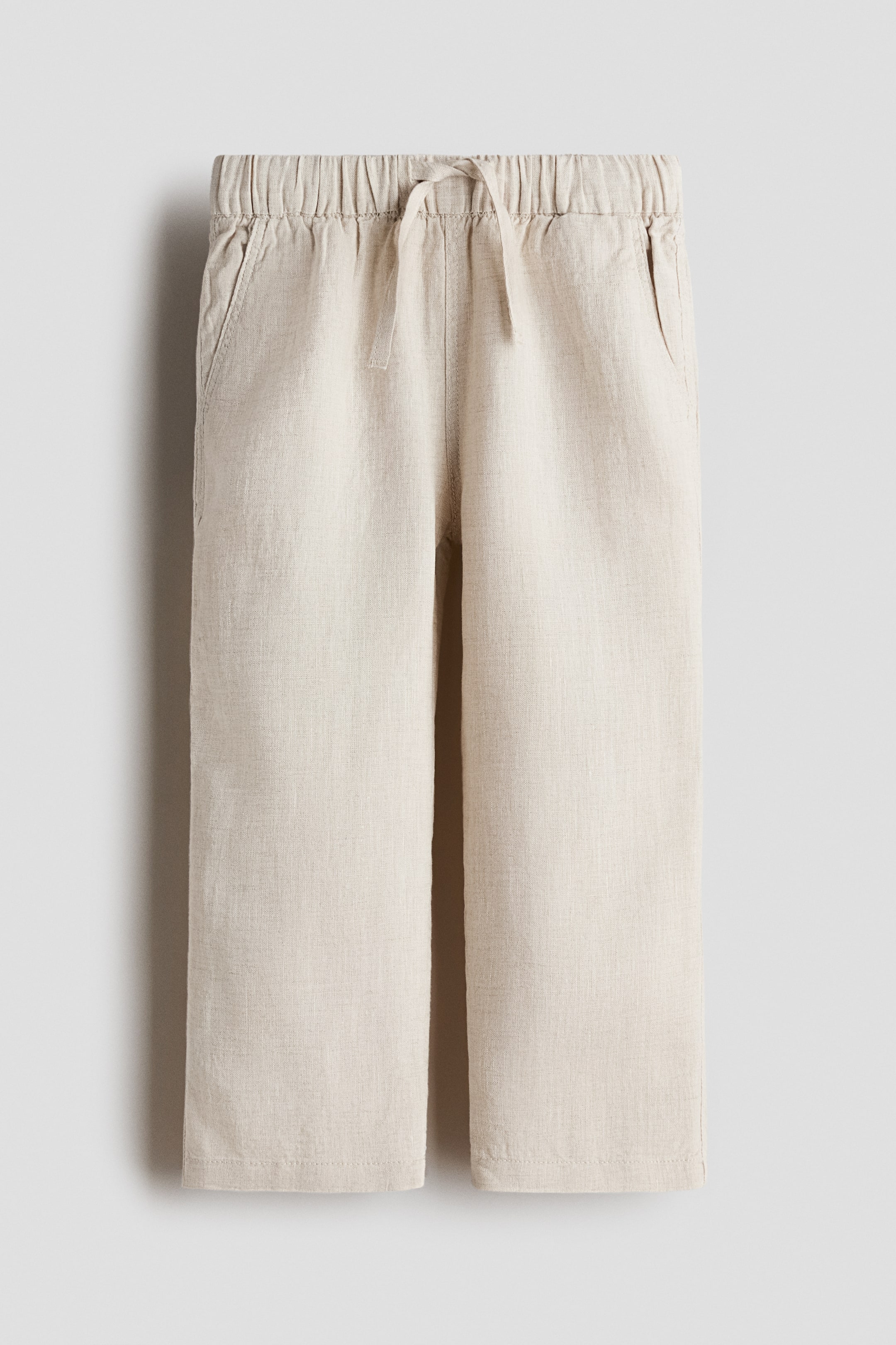 Agrandir l'image: Un pantalon beige clair texturé est présenté en vue de face, doté d'une taille élastiquée avec un cordon de serrage central. Des poches fendues sont visibles sur les hanches, et le pantalon est conçu avec des jambes droites.