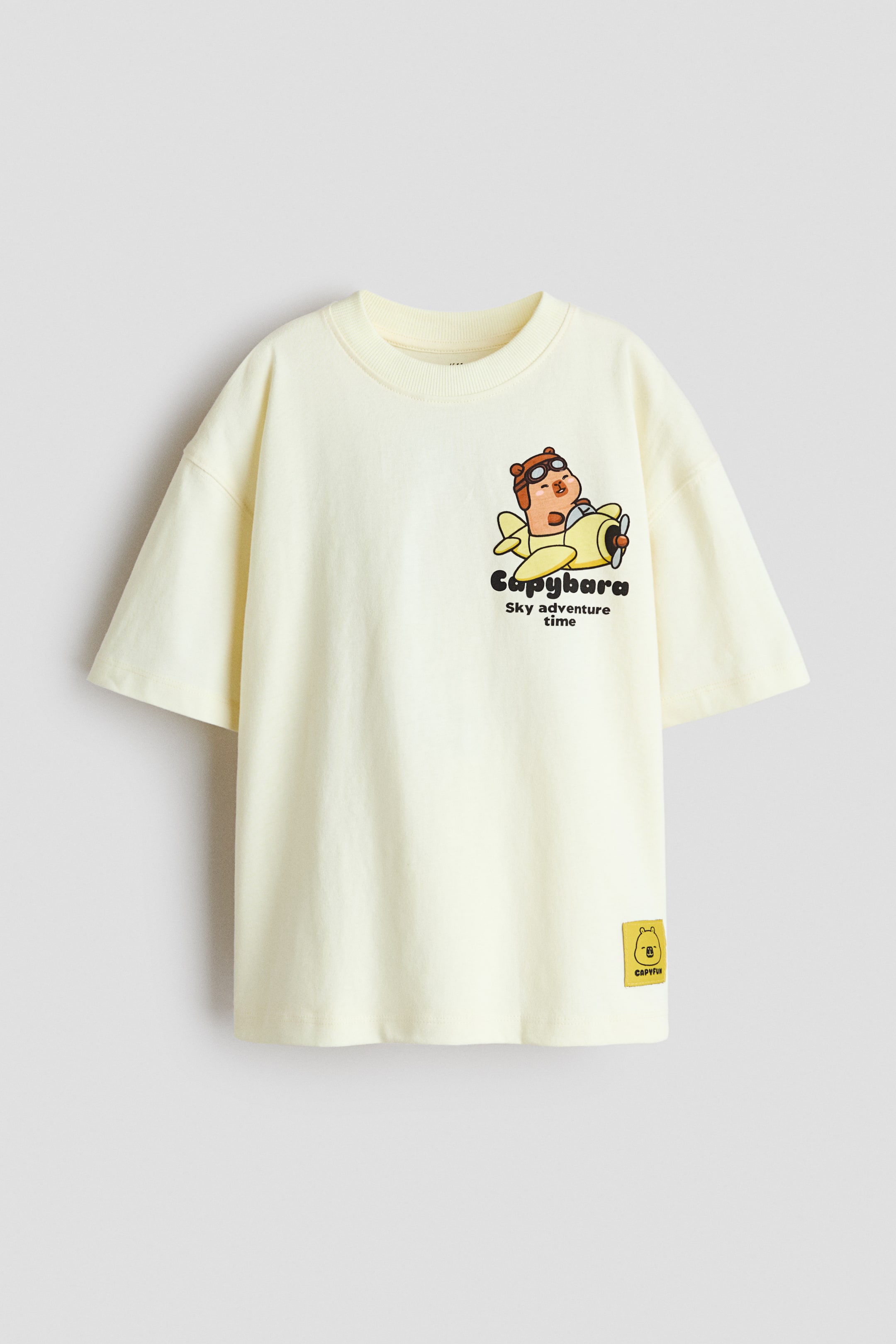 Zobacz większy obraz: Luźno dopasowany T-shirt z miękkiej bawełnianej dzianiny z nadrukowanymi motywami prezentuje widok z przodu, posiadając okrągły dekolt z prążkowanym wykończeniem i obniżone ramiona. Główny nadruk przedstawia kreskówkowego kapibarę w okularach pilota, siedzącego w żółtym dwupłatowcu, z towarzyszącym tekstem "Capybara Sky adventure time", a na dolnym prawym brzegu widoczna jest mała żółta metka marki.